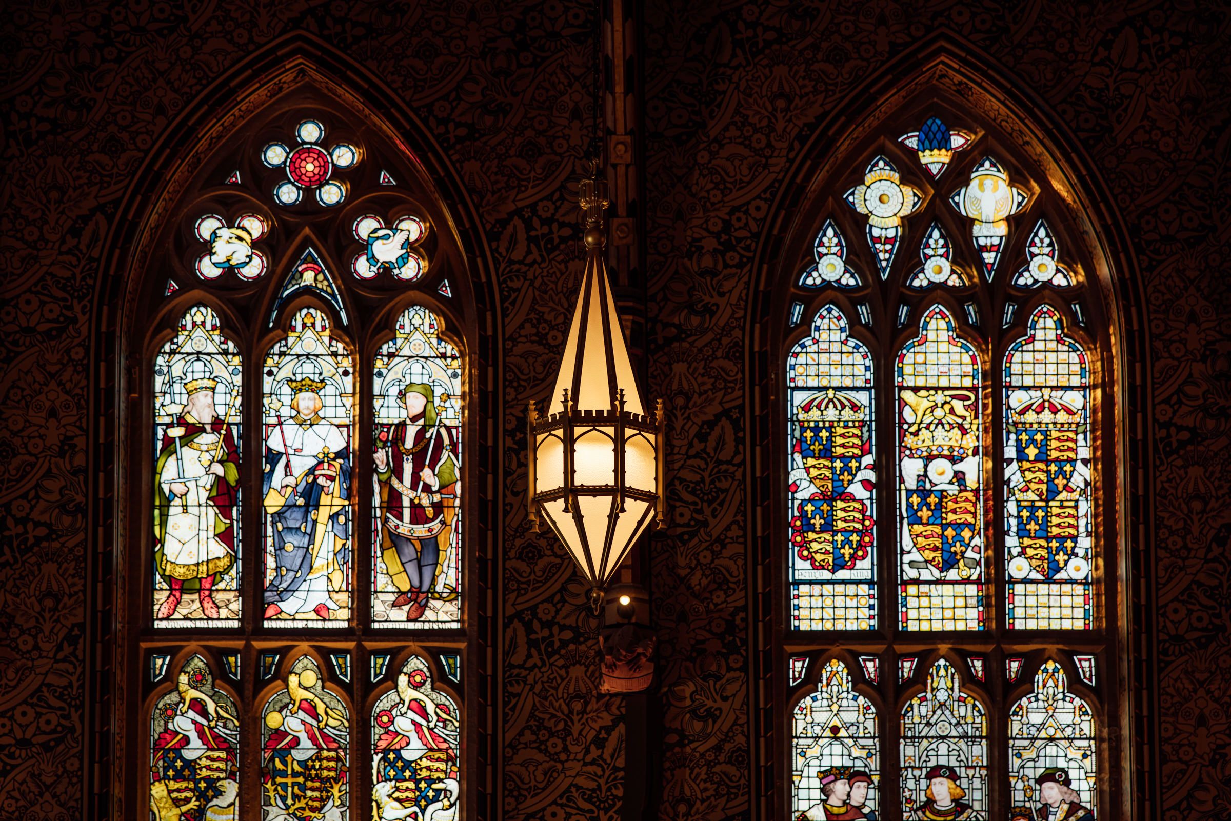 Two Gothic arched stained glass windows with robed figures and heraldic shields flank a hanging brass lantern in a dark paneled hall.