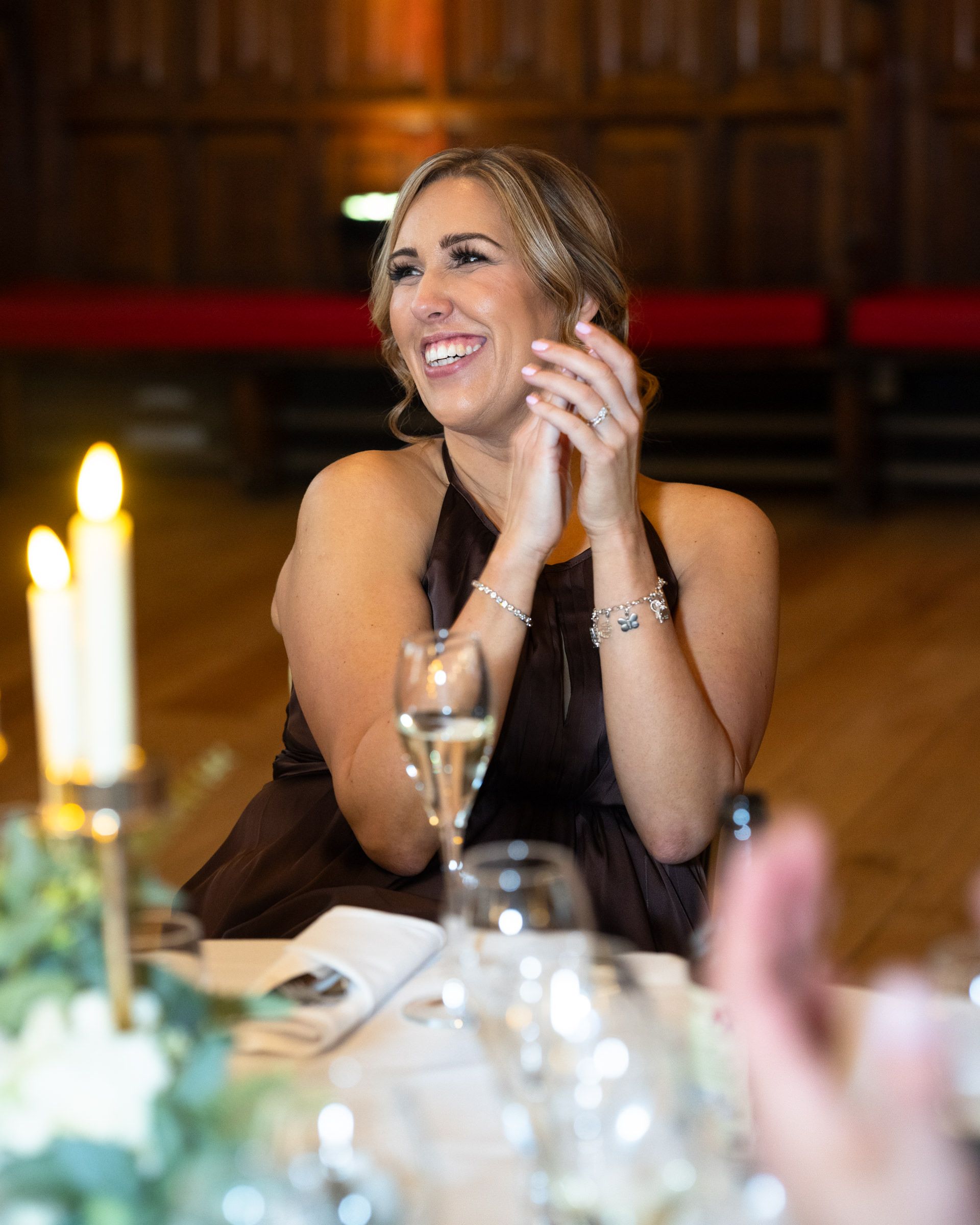 Smiling woman in a dark halter dress applauds at a candlelit reception table with champagne glasses and flowers in a wood-paneled hall.