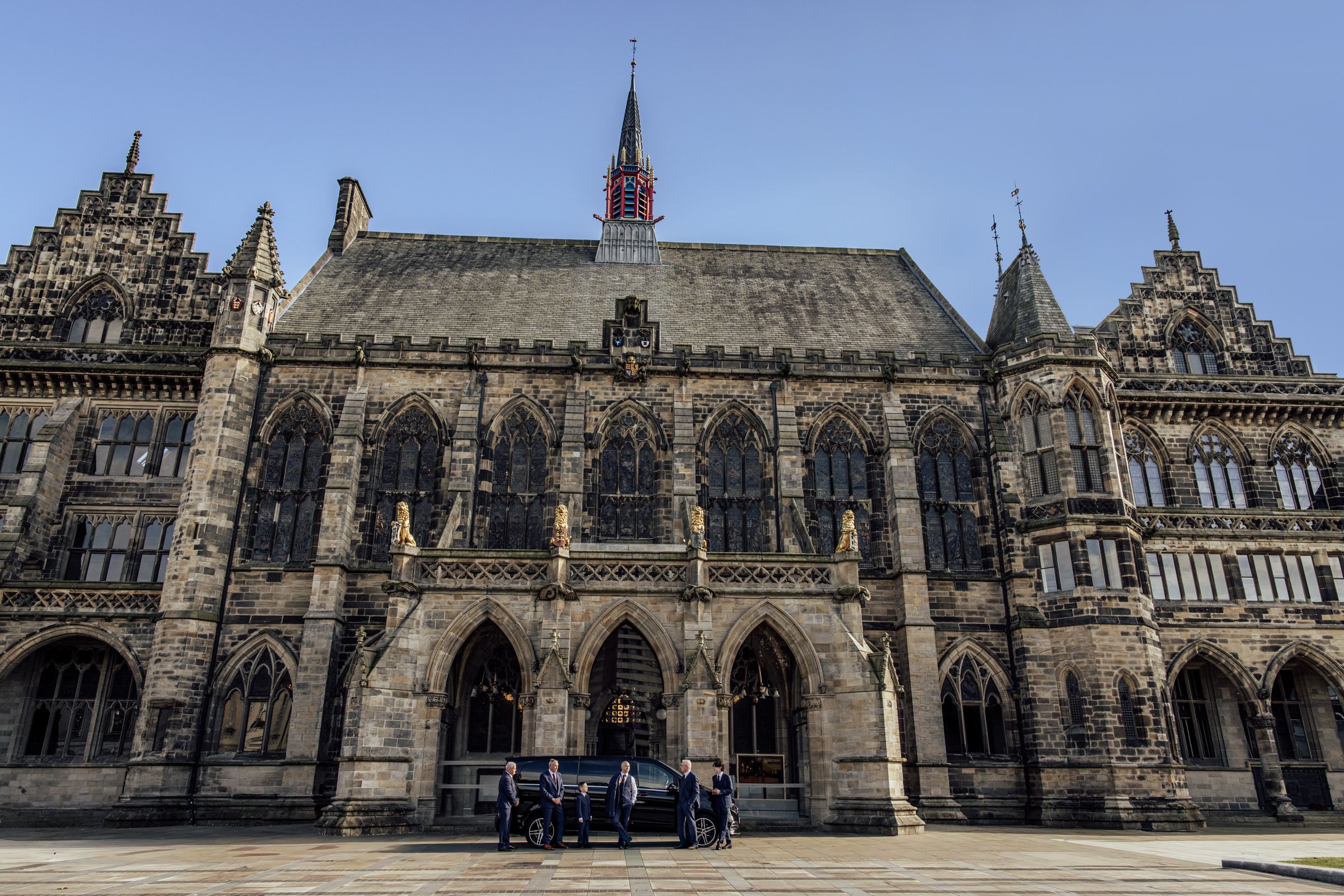 Gothic Revival town hall with ornate stone facade, pointed arch windows and a central spire, with people gathered at the entrance under a bl