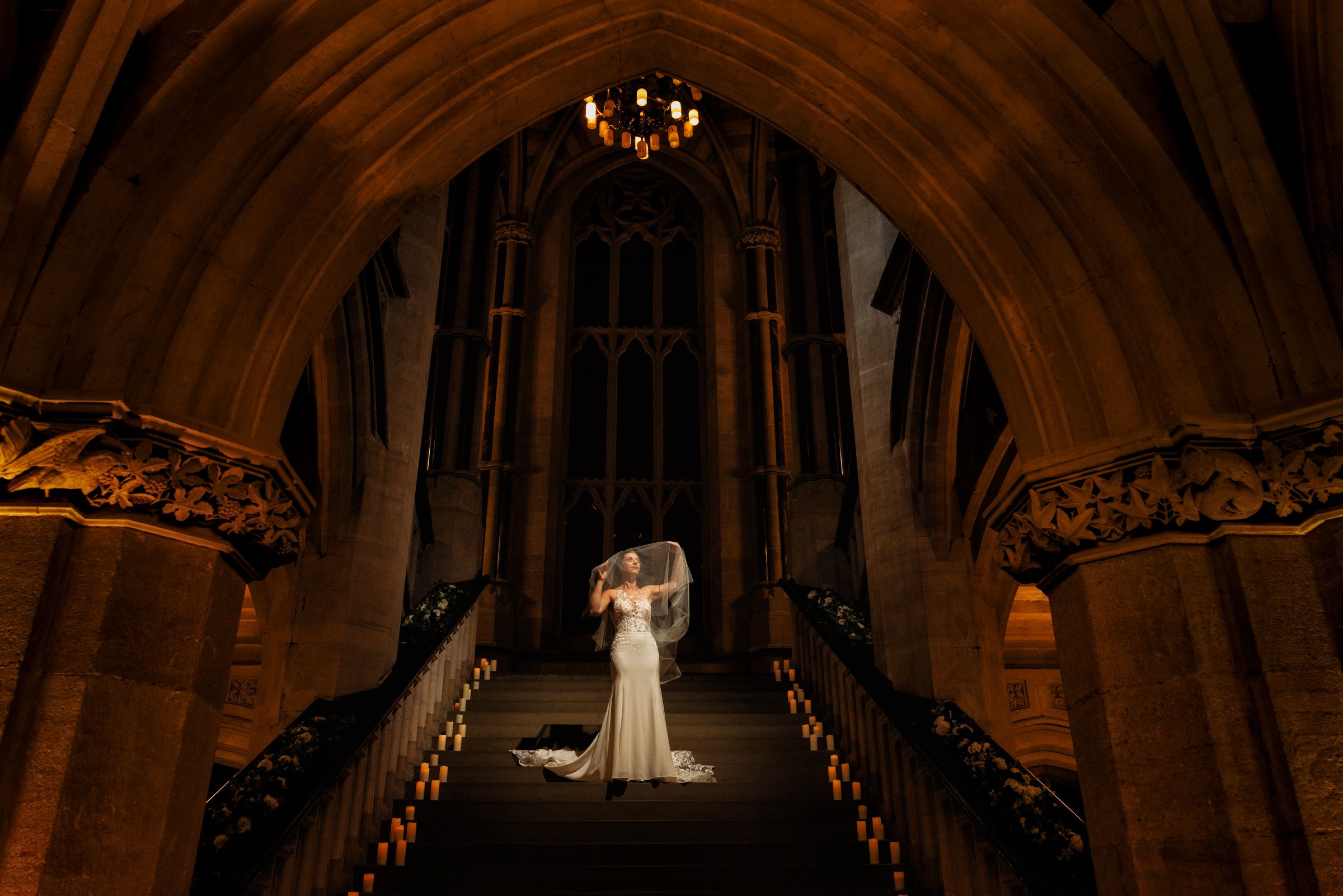 Bride in a white gown and flowing veil stands on grand stone steps beneath a gothic arch and glowing chandelier in a historic cathedral.