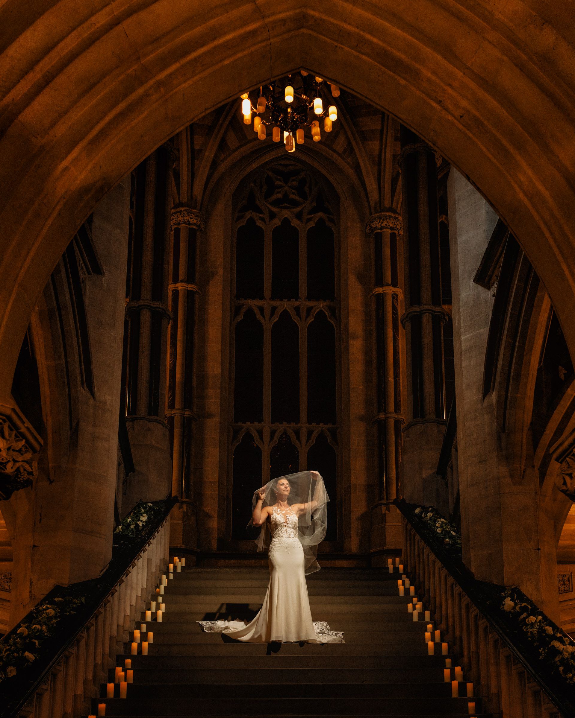 Bride in a white gown and flowing veil stands on grand stone steps lined with candles beneath a Gothic arch and glowing chandelier.