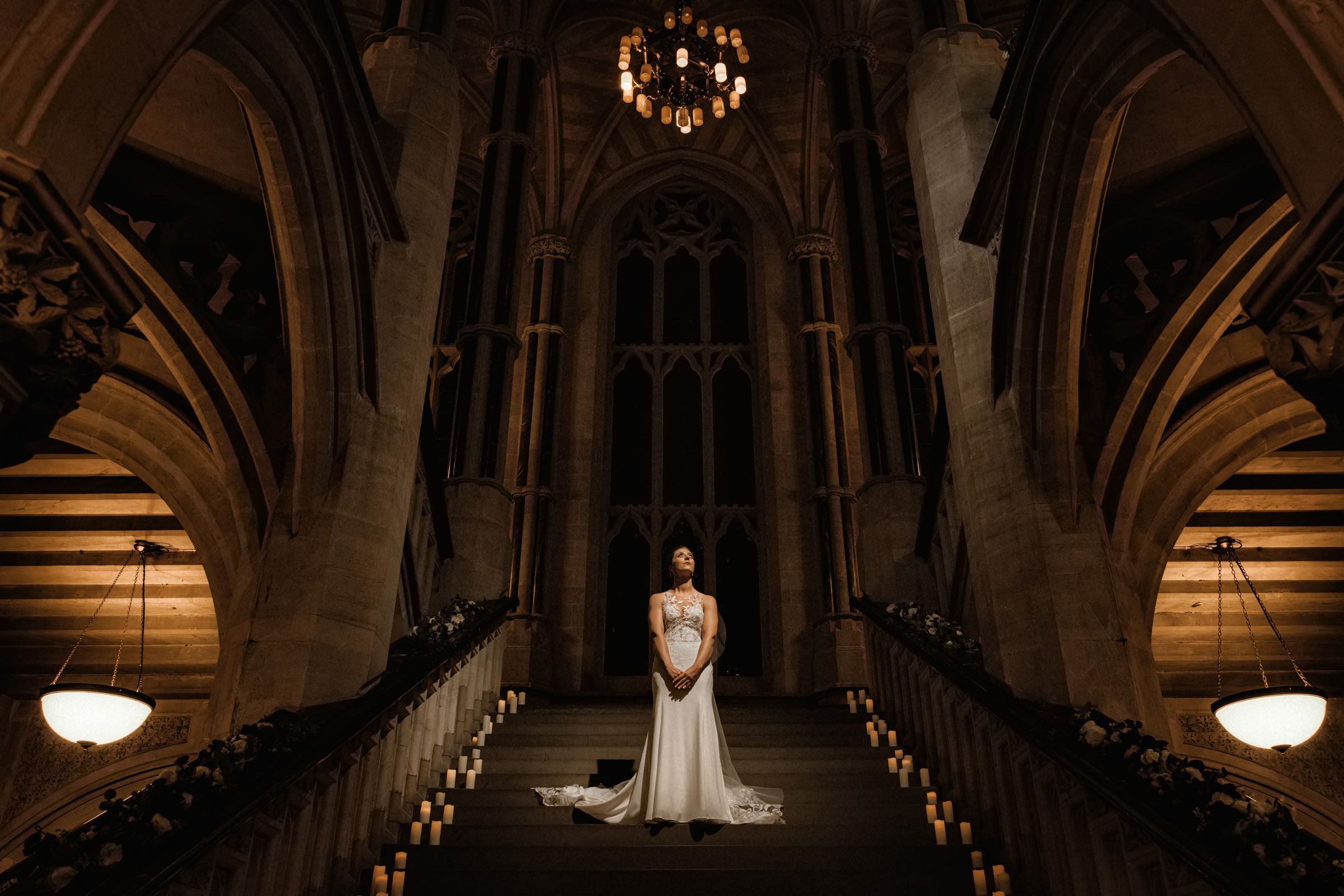 Bride in a white gown stands at the end of a candlelit aisle inside a grand Gothic cathedral with soaring arches and a glowing chandelier.