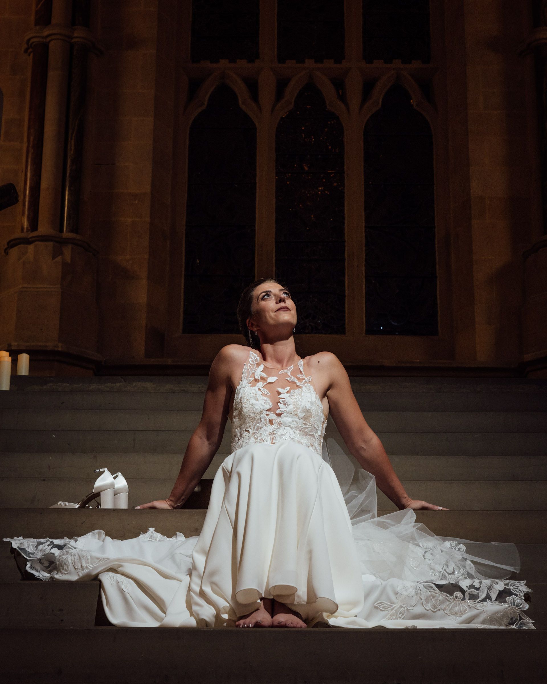 Barefoot bride in a white lace wedding gown sits on stone church steps, gazing upward beneath a Gothic window, with her shoes beside her.