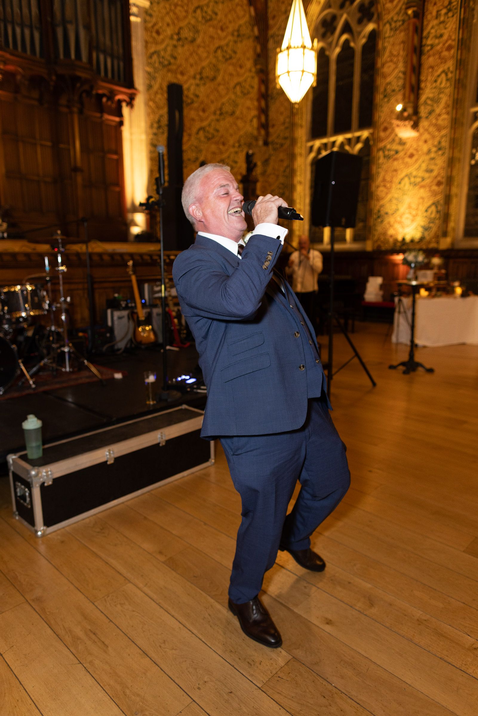 Man in a blue suit singing into a microphone on a wooden dance floor in a historic hall with a drum kit and band equipment behind him.
