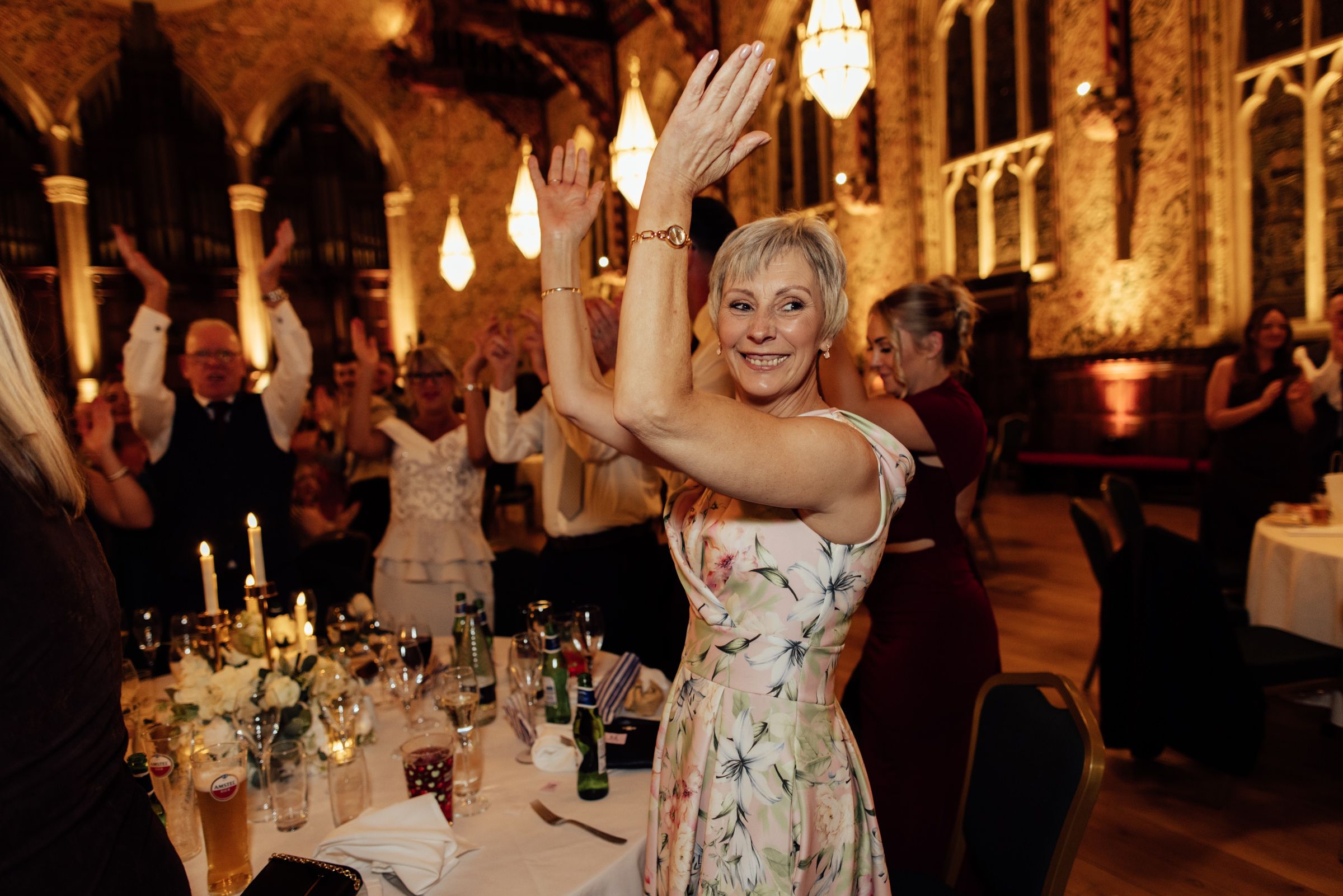 Smiling woman in a floral dress raises her arms and dances at a candlelit wedding reception as guests cheer in a historic hall.