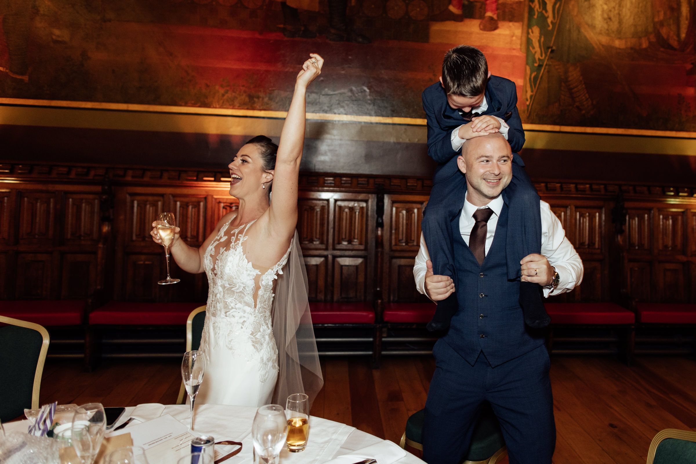 Bride in a lace gown and veil raises her fist and a glass in joy as a groom in a navy suit carries a man on his shoulders.