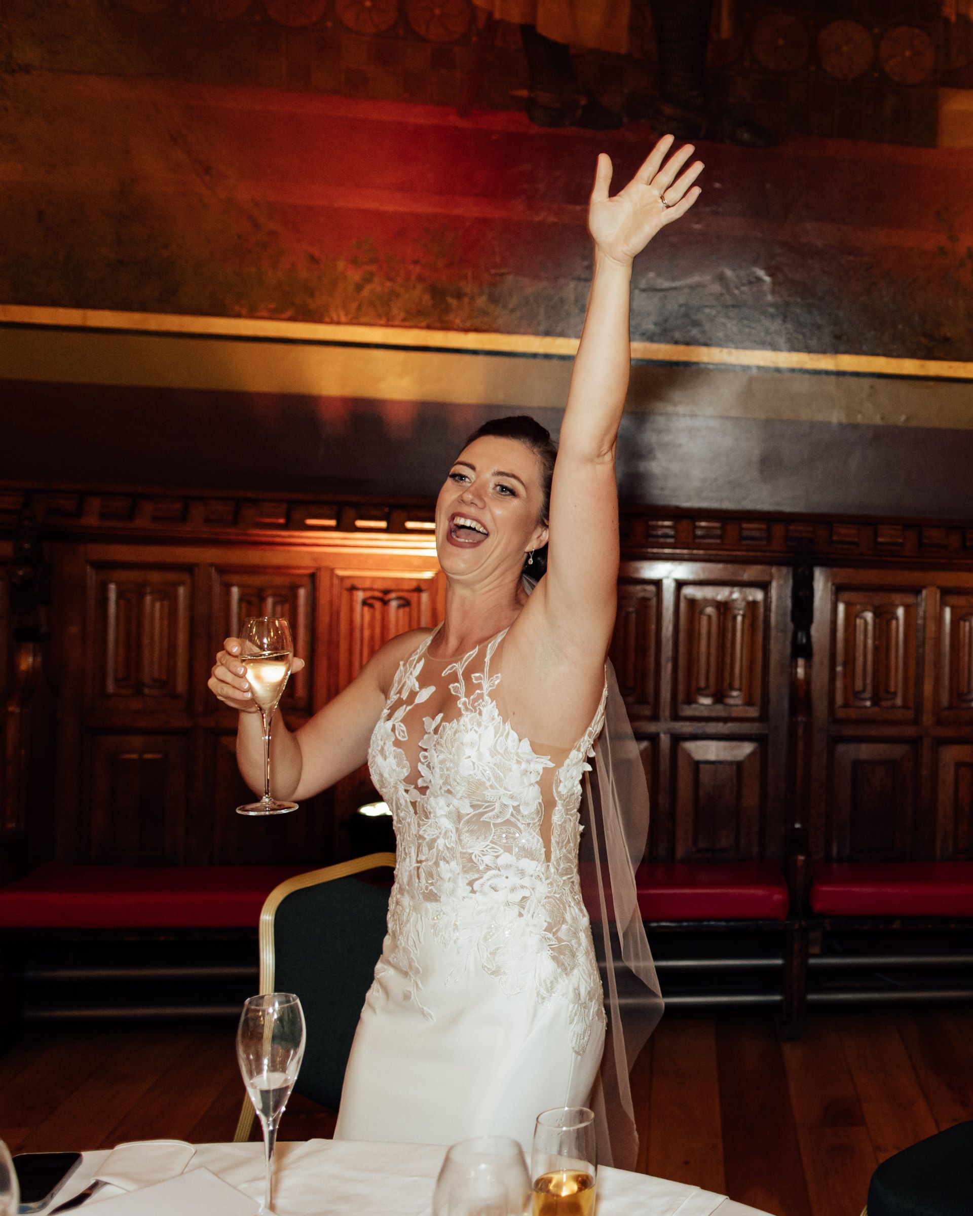 A joyful bride in a lace wedding gown raises her arm and a champagne glass during a celebratory toast at a wedding reception.