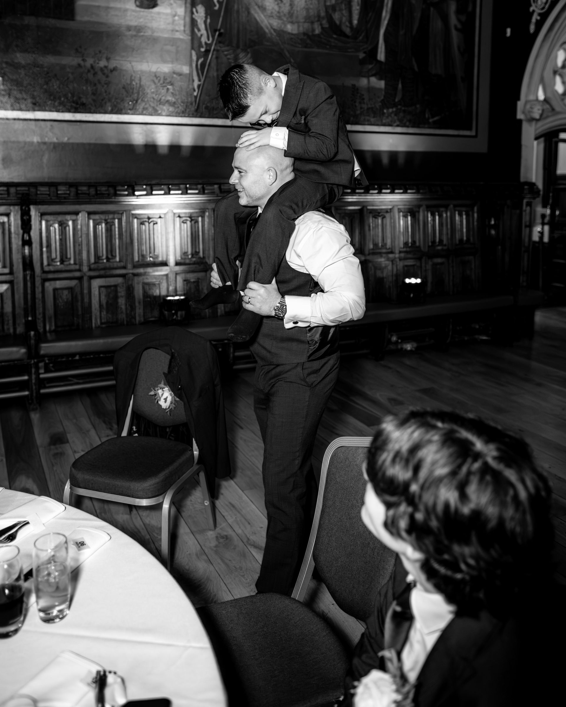 Black and white photo of a groomsman lifting another man on his shoulders in a wedding reception hall as a guest looks on.