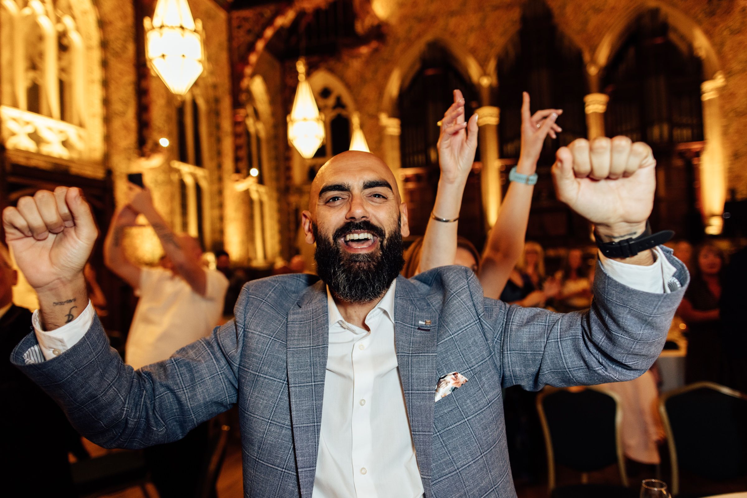 Bearded man in a gray blazer cheers with raised fists and a big smile at a lively event inside a Gothic-style venue with arched windows.