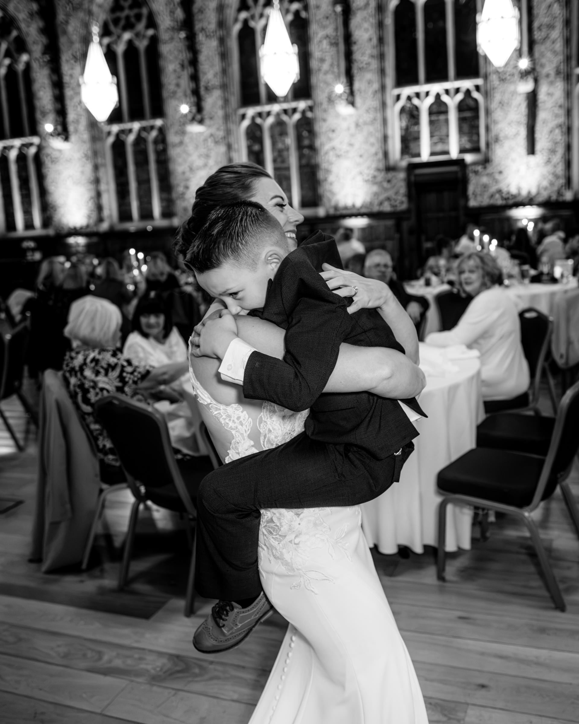 Black and white photo of a bride in a lace wedding gown embracing a young boy in a suit during a joyful reception in a grand hall.