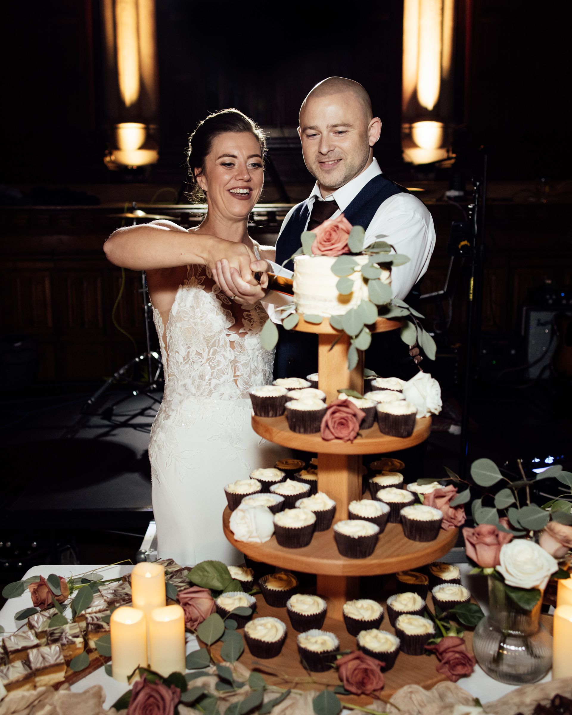 A bride and groom smile as they cut a cake together beside a tiered cupcake stand decorated with roses, greenery and glowing candles.