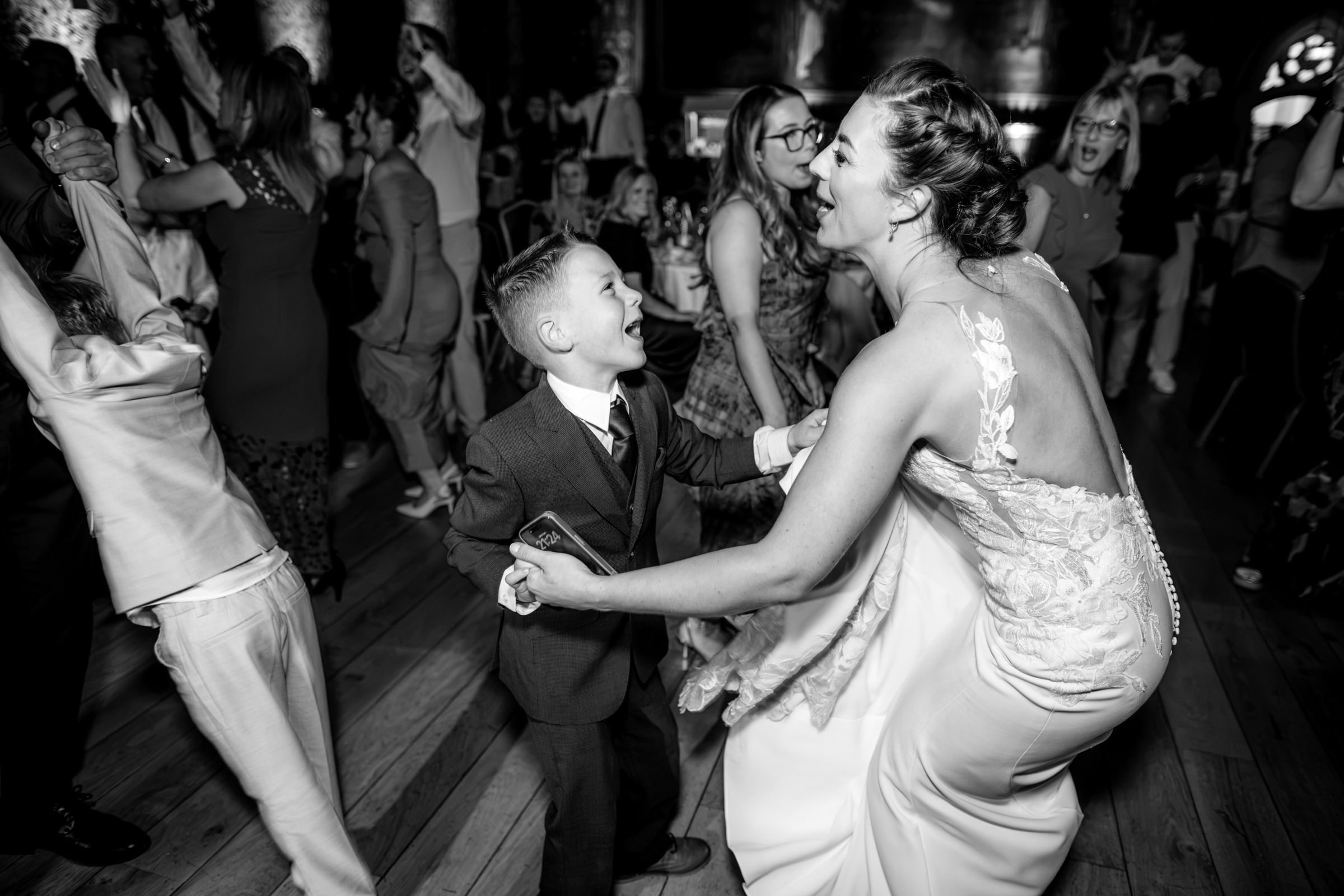 Bride in a lace wedding gown dances joyfully with a young boy in a suit on a crowded wedding reception dance floor in black and white.