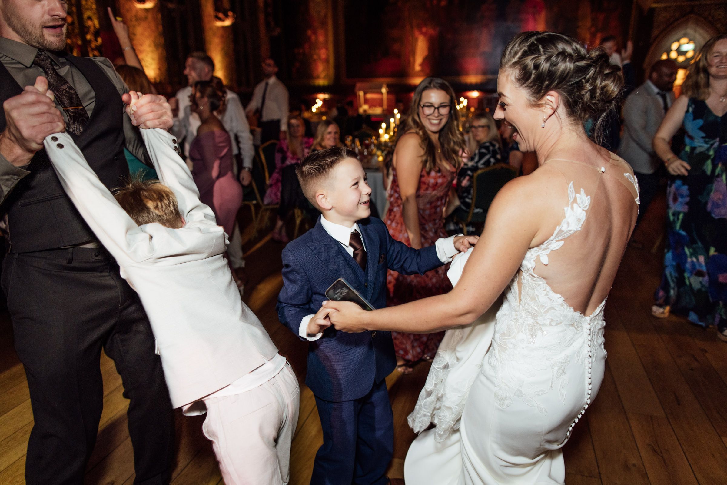 Bride in a lace wedding gown dances with a young boy in a navy suit as guests celebrate on the dance floor at a wedding reception.