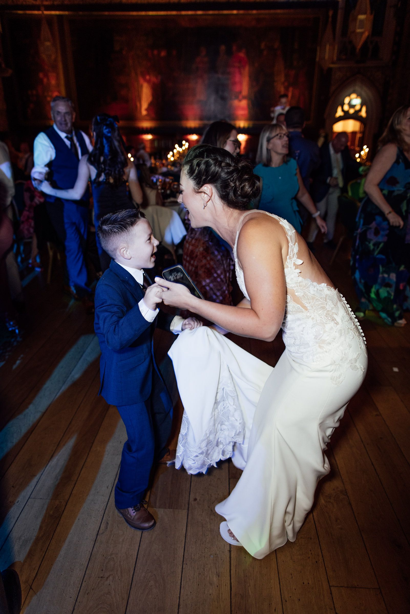 A bride in a lace wedding dress dances joyfully with a young boy in a navy suit while guests mingle in a warmly lit reception hall.