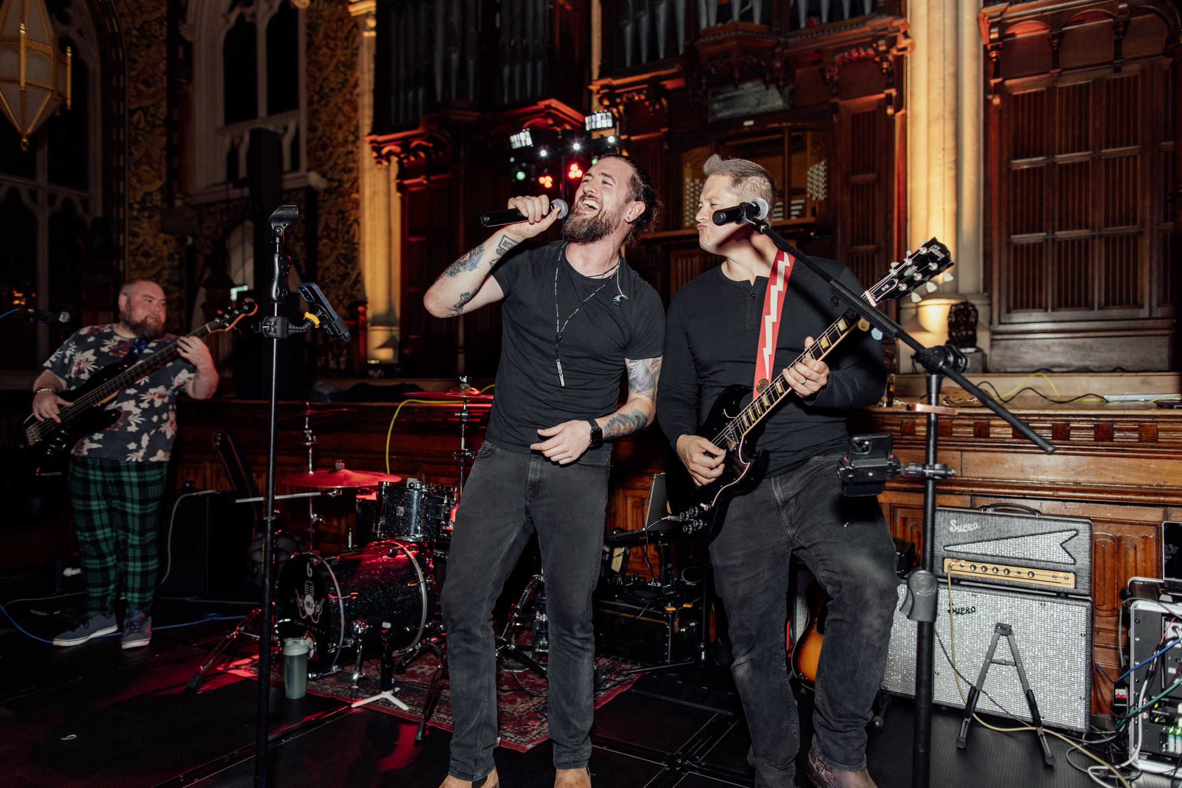 Bearded singer with tattoos performs live on stage with a guitarist and drummer in a church-style venue with wood paneling.
