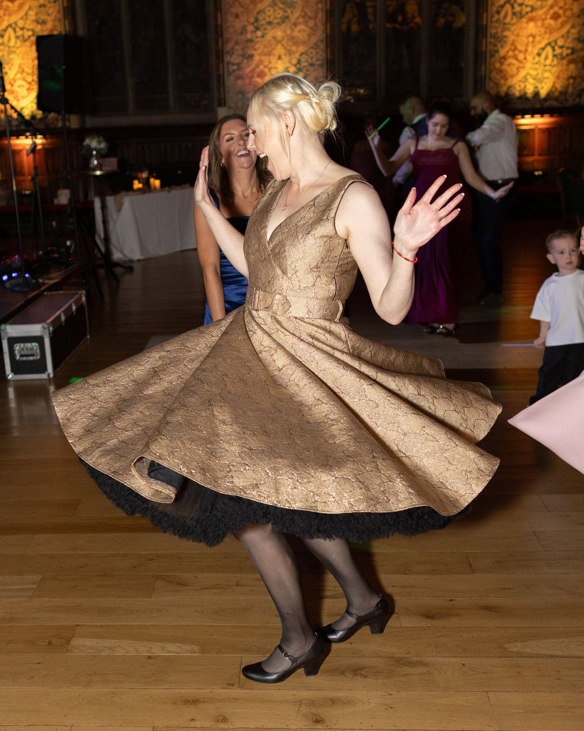 Blonde woman twirls in a gold brocade dress with black petticoat on a wooden dance floor at a lively evening event.