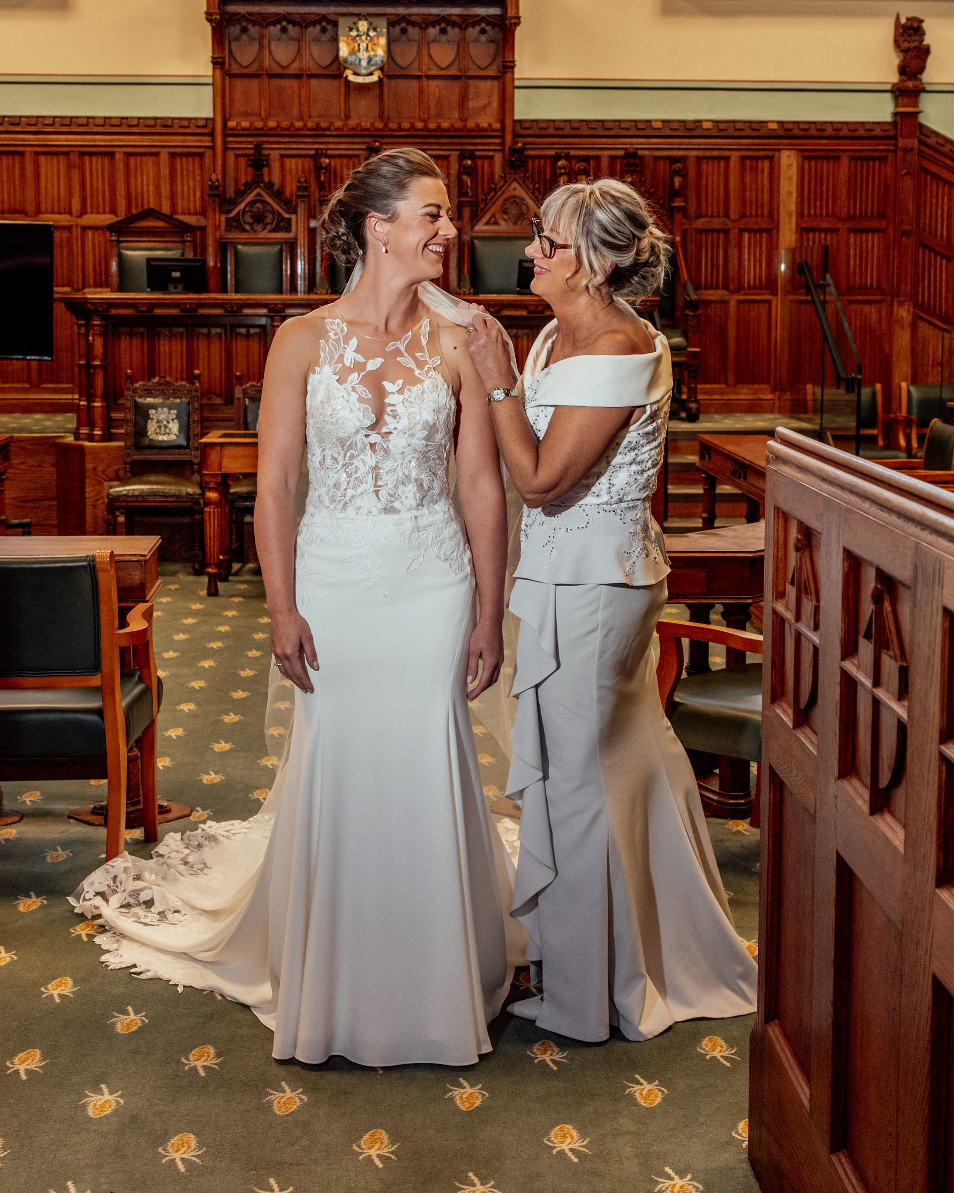 A bride in a lace-bodice white gown shares a joyful moment with a woman in a gray dress in a wood-paneled room with patterned carpet.