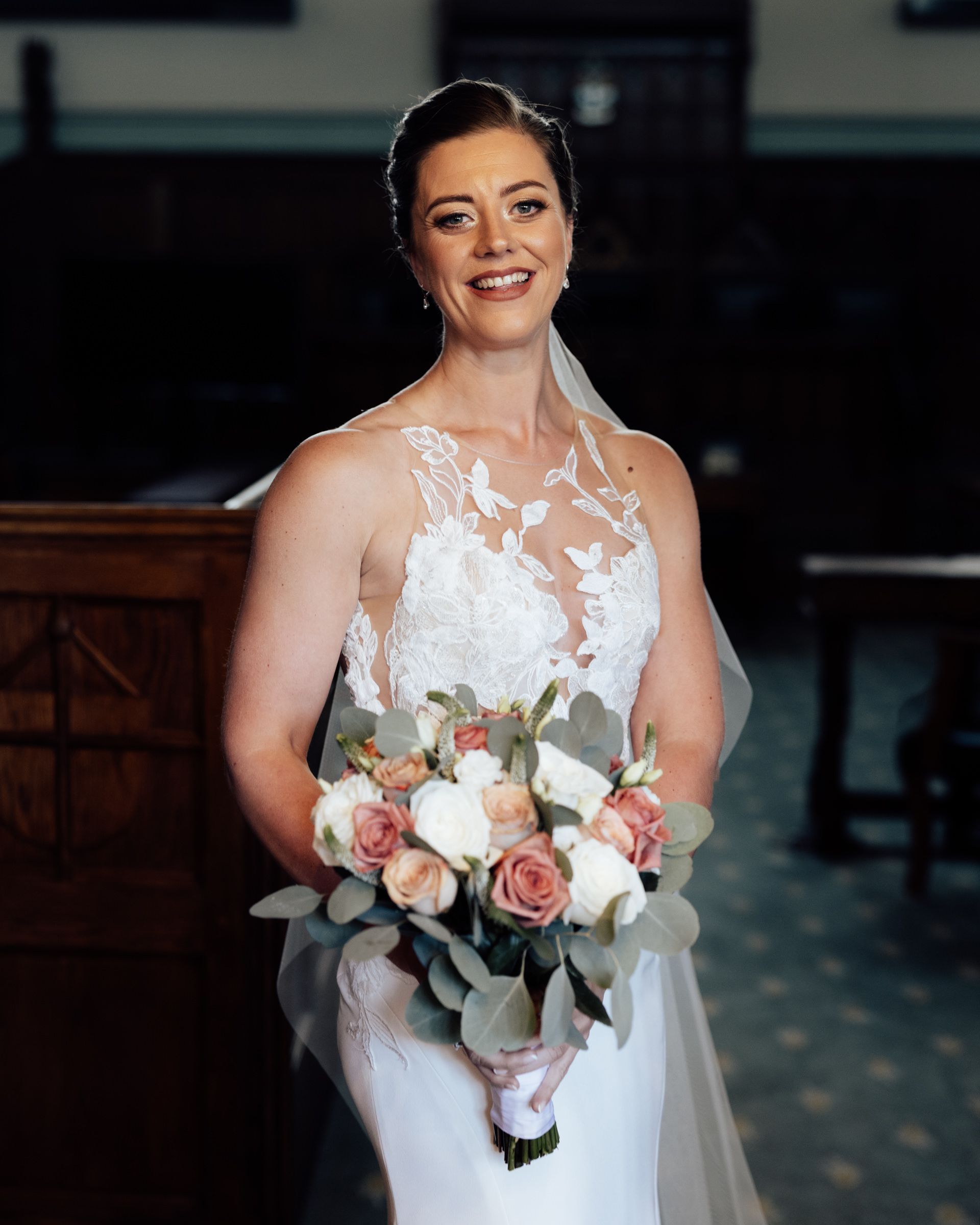 Smiling bride in a white lace sleeveless wedding gown and veil holding a bouquet of pink and white roses with eucalyptus leaves.
