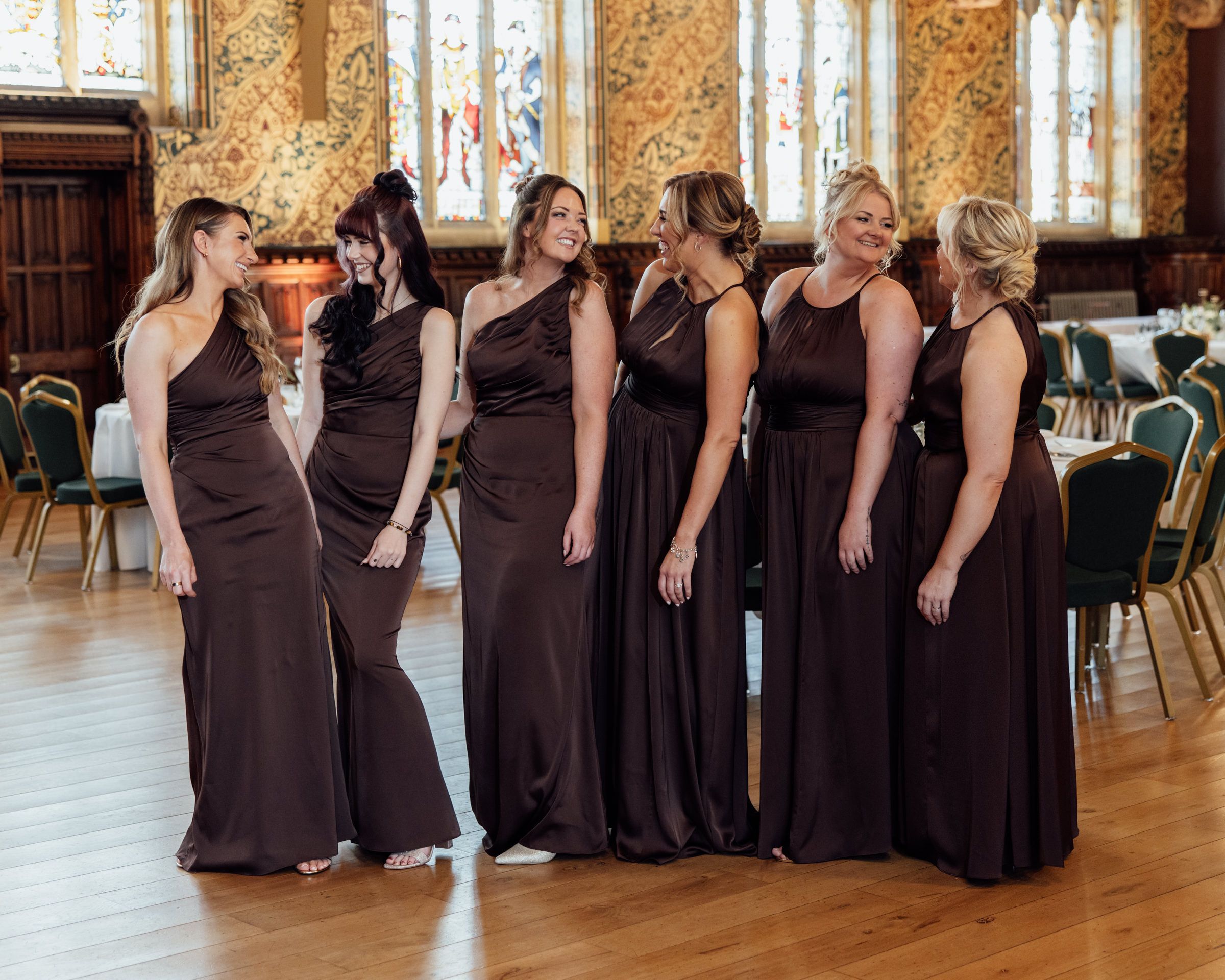 Six bridesmaids in matching chocolate brown satin dresses stand in a row, smiling and laughing in an elegant wedding venue with wood floors.