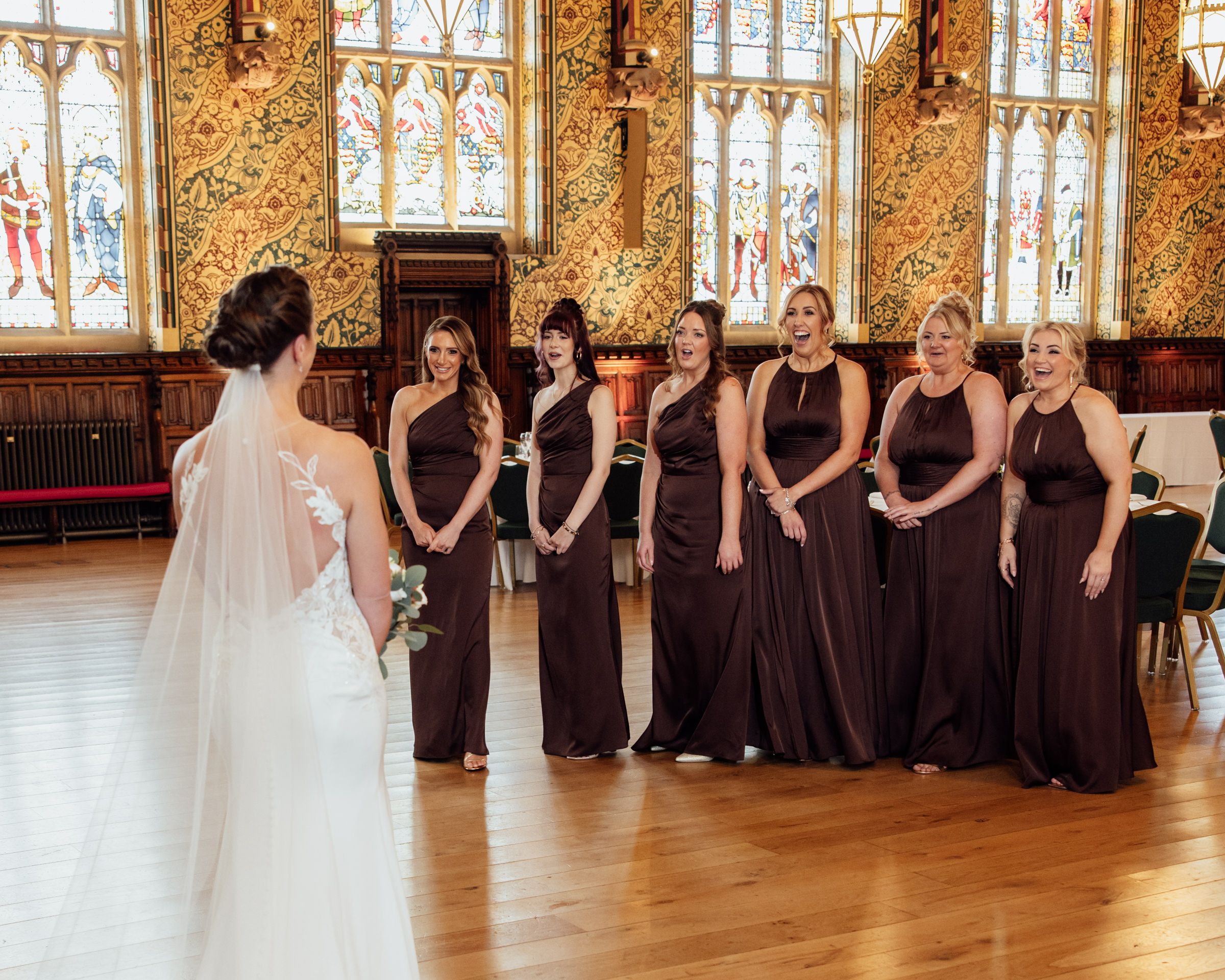 Bride in a lace gown and long veil faces six bridesmaids in brown dresses in an ornate hall with stained glass windows.