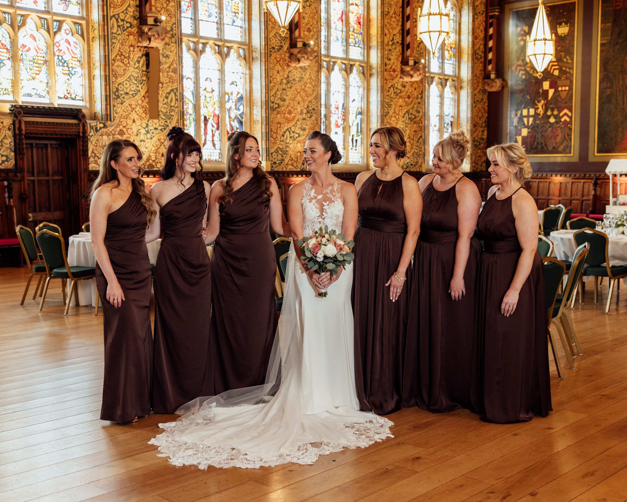 Bride in a lace wedding gown holding a bouquet, surrounded by seven bridesmaids in chocolate brown dresses in an elegant ballroom venue.