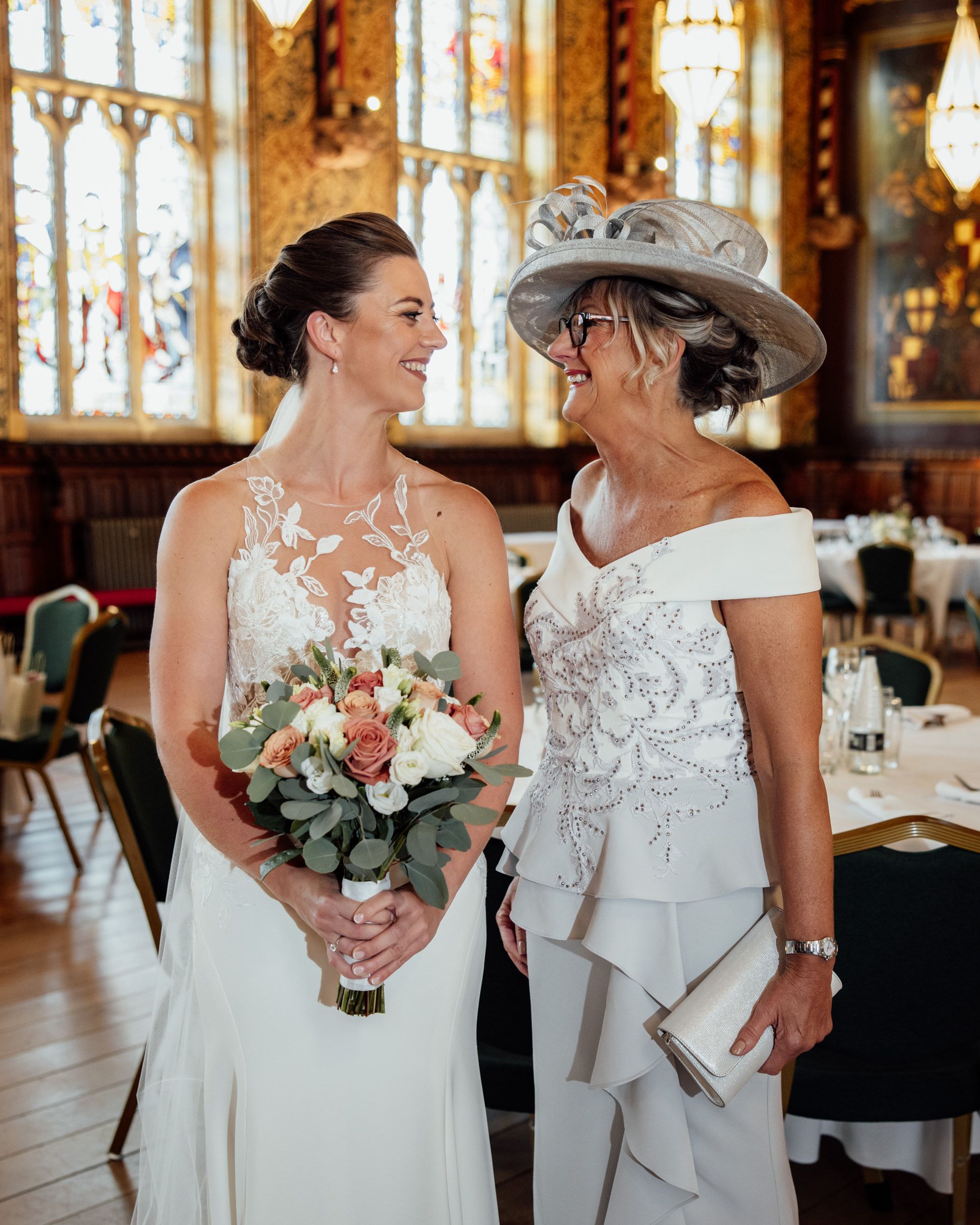 Smiling bride in a lace gown holding a pink and white bouquet shares a joyful moment with a woman in an ivory dress and fascinator hat.