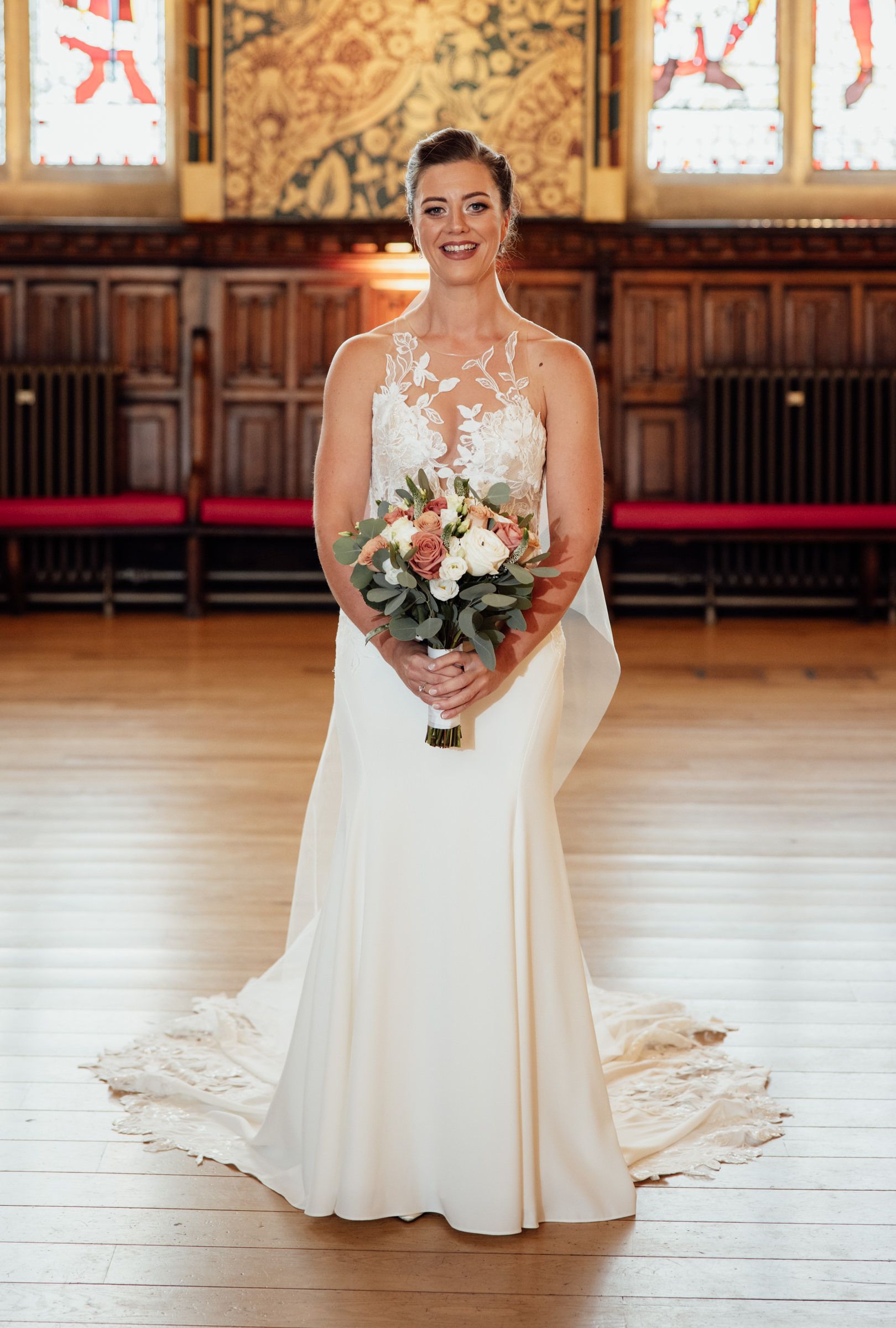 Smiling bride in an elegant lace and satin wedding gown holding a pink and white bouquet in a grand hall with wood floors.