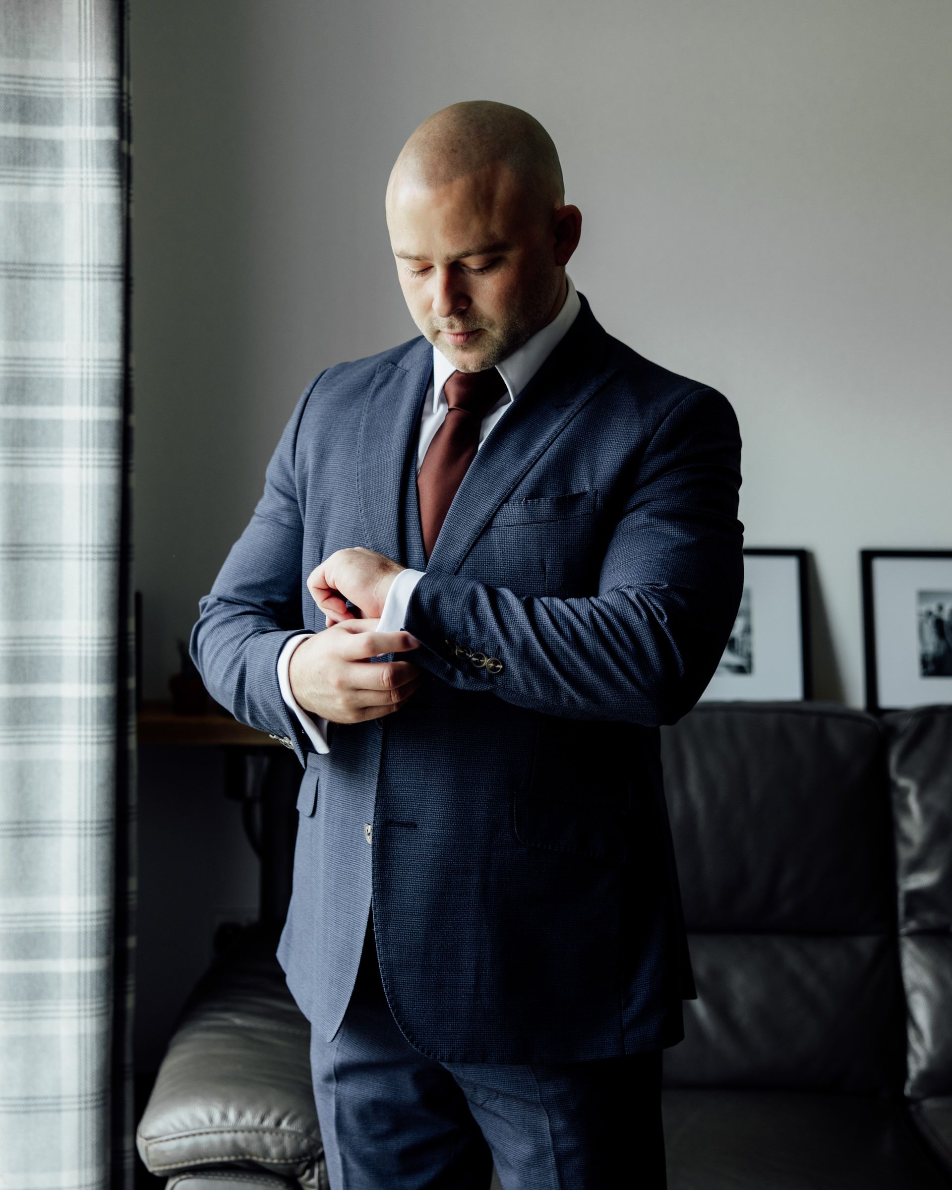 Groom in a navy suit and burgundy tie adjusts his shirt cuff by a window in a softly lit room with a leather sofa.