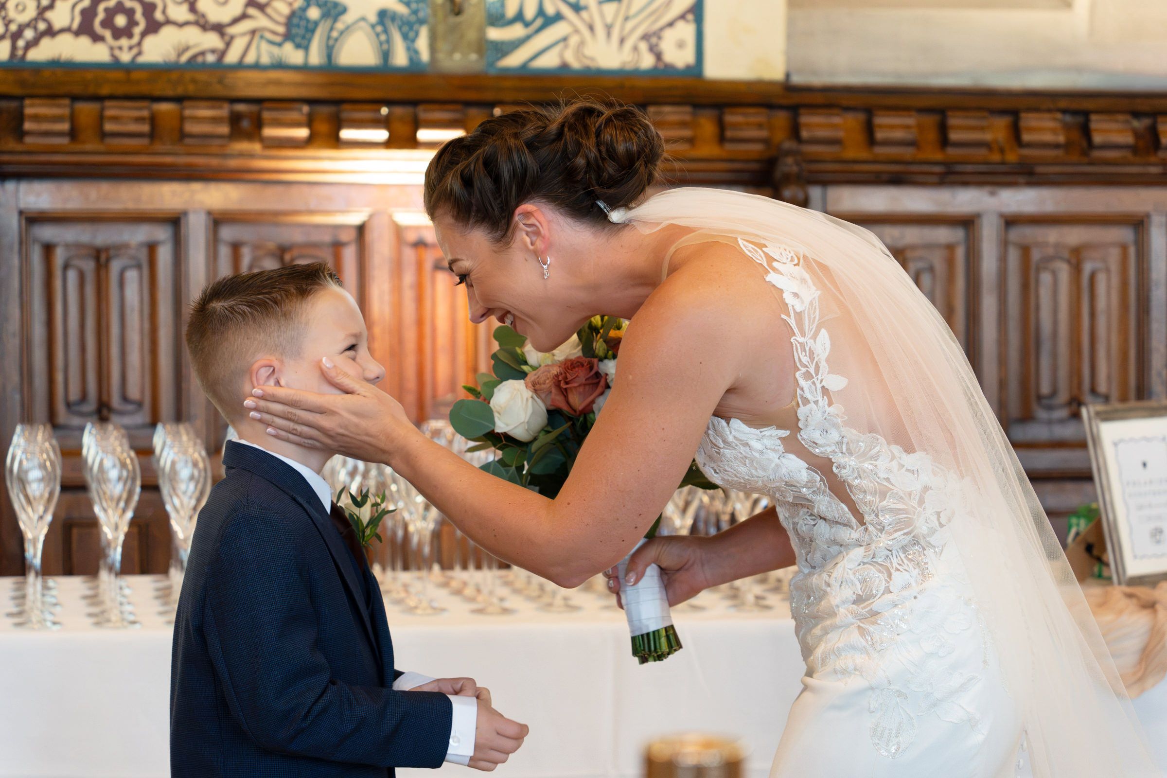 Smiling bride in a lace wedding gown and veil holding a bouquet gently touches the cheek of a young boy in a navy suit at the reception.
