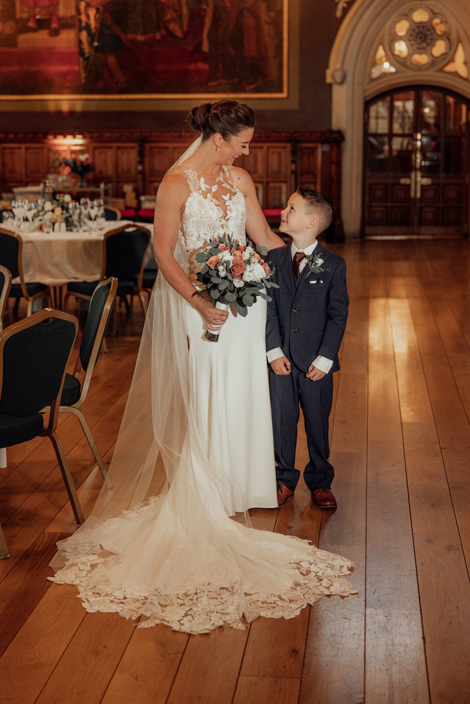 Bride in a lace wedding dress with a long veil holds a bouquet and smiles down at a young boy in a navy suit in a historic hall.