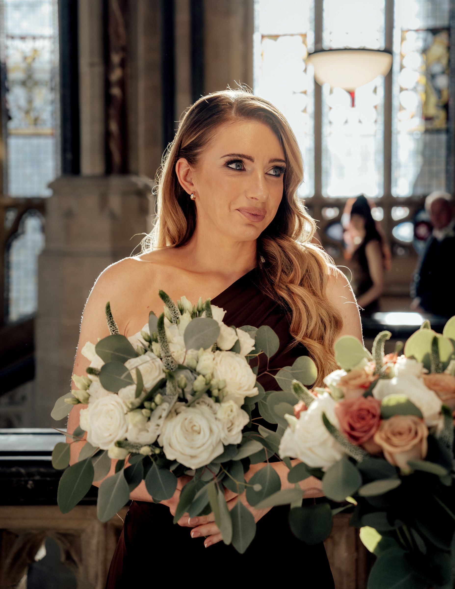 Bridesmaid in a dark burgundy dress holds a bouquet of white and blush roses with eucalyptus inside a church during a wedding ceremony.
