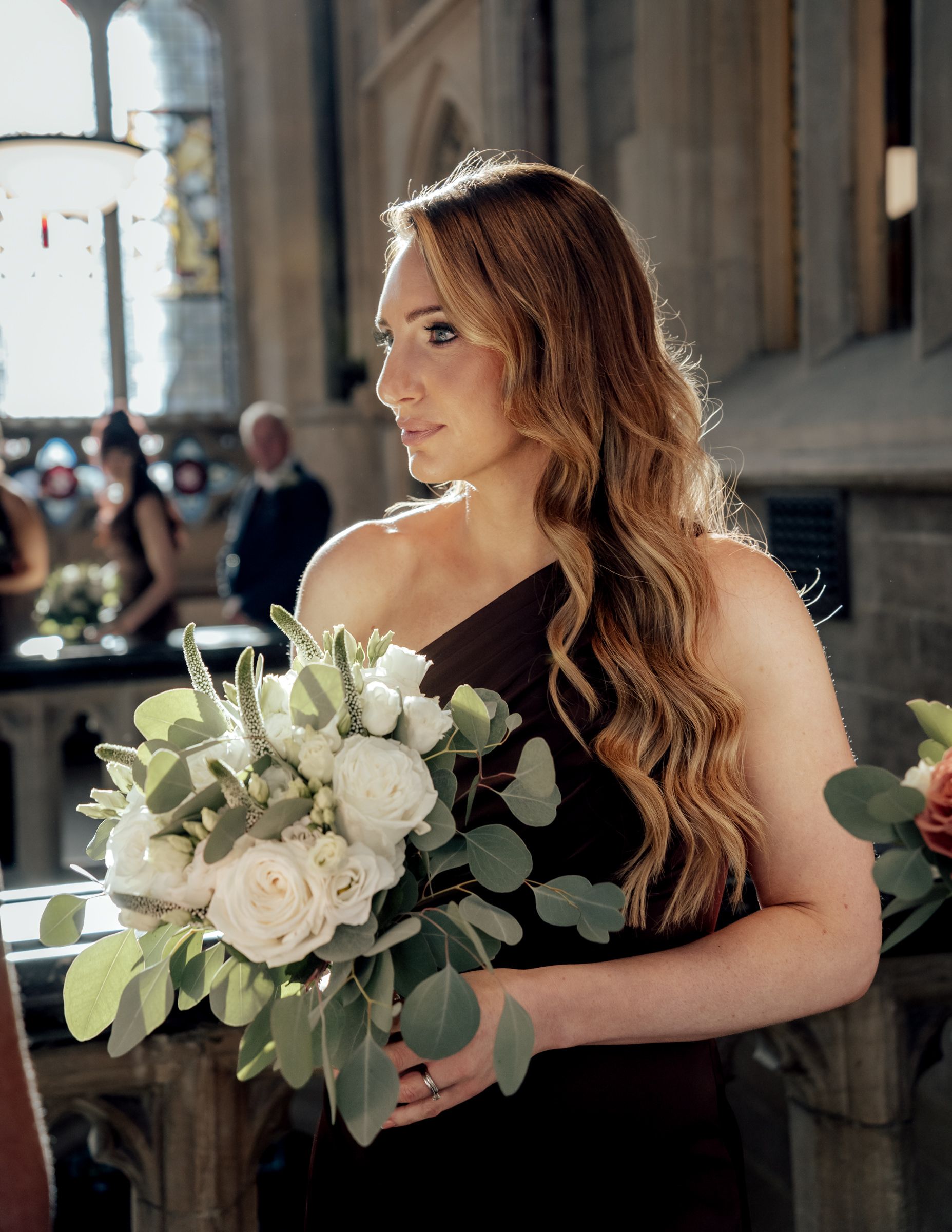 Bridesmaid in a dark one-shoulder dress with long wavy auburn hair holds a white rose and eucalyptus bouquet inside a stone church.