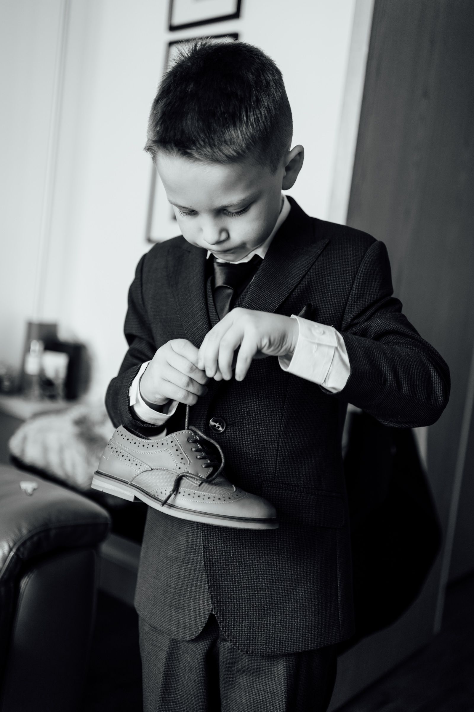 Black and white photo of a young boy in a suit and tie carefully tying the laces of a brogue shoe indoors.
