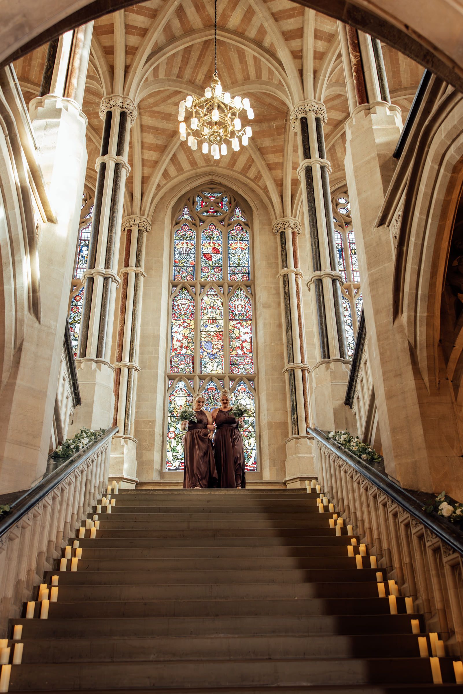 Two bridesmaids in burgundy dresses stand atop a grand staircase lined with candles beneath a stained glass window and chandelier.