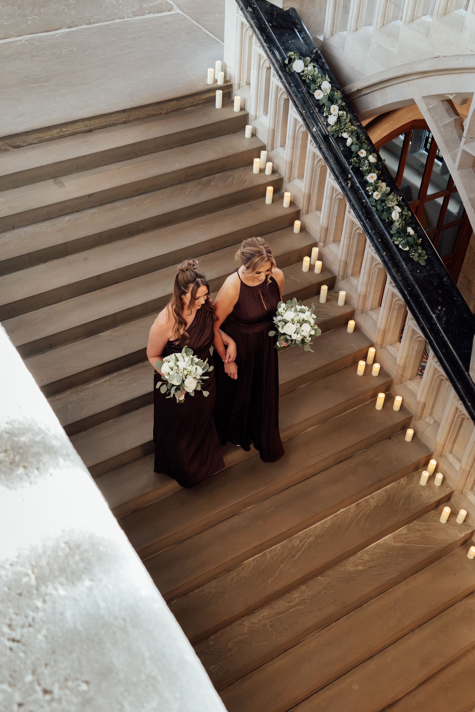 Two bridesmaids in dark dresses hold white bouquets while walking arm in arm down a candlelit staircase with floral garland.