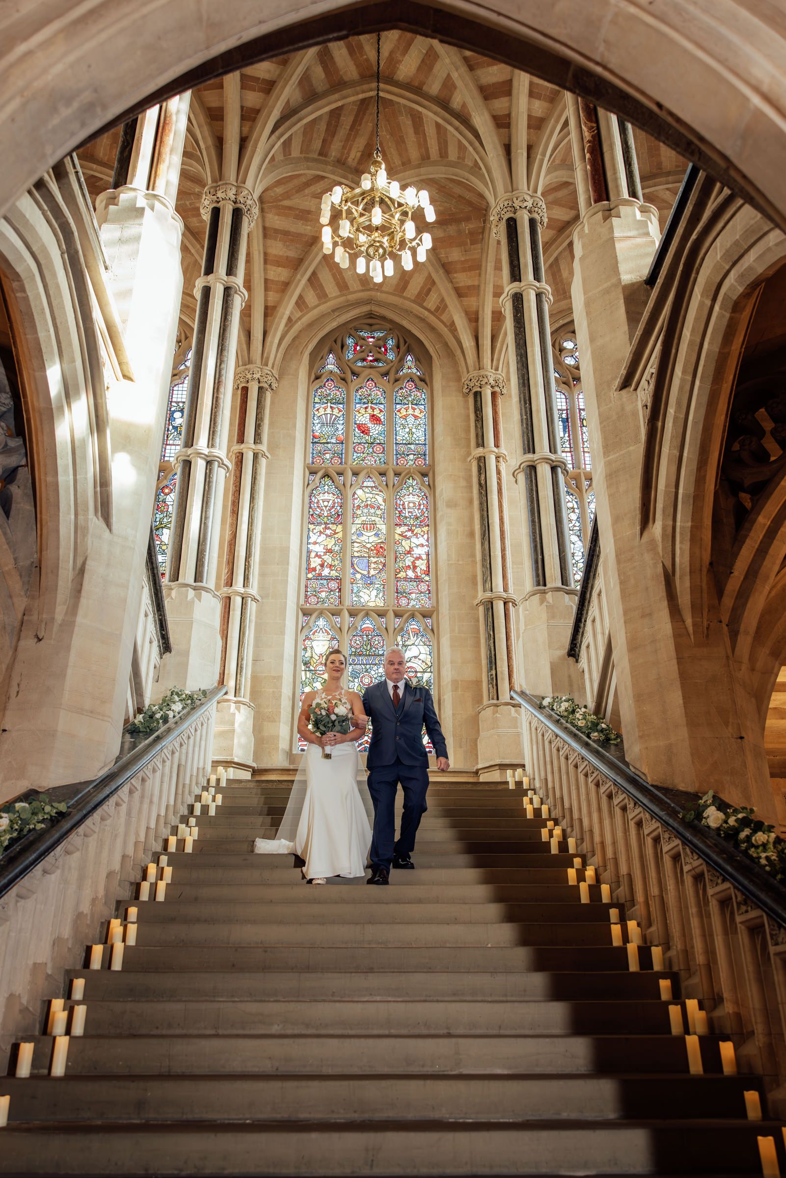 Bride in a white gown and groom in a navy suit walk down a grand Gothic stone staircase beneath a chandelier and stained glass window.