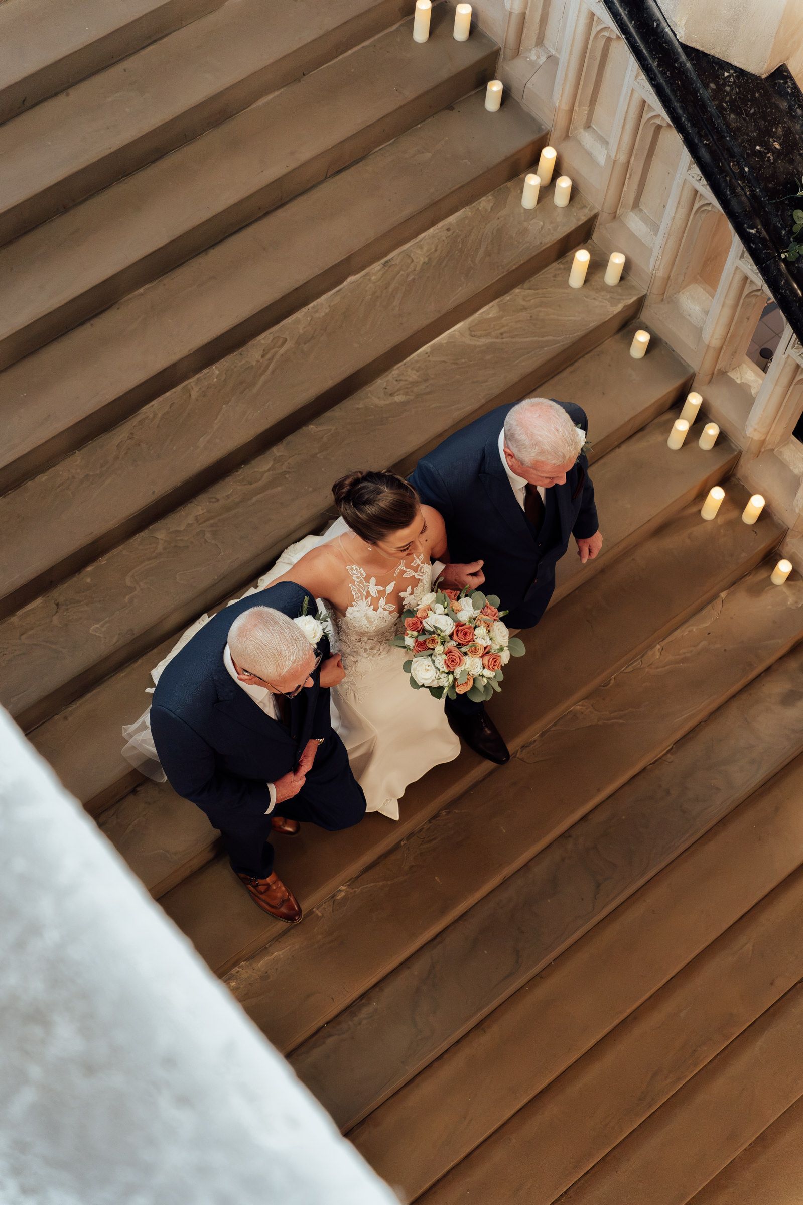 Overhead view of a bride in a white gown holding a bouquet while walking down a wide staircase lined with candles between two men in suits.