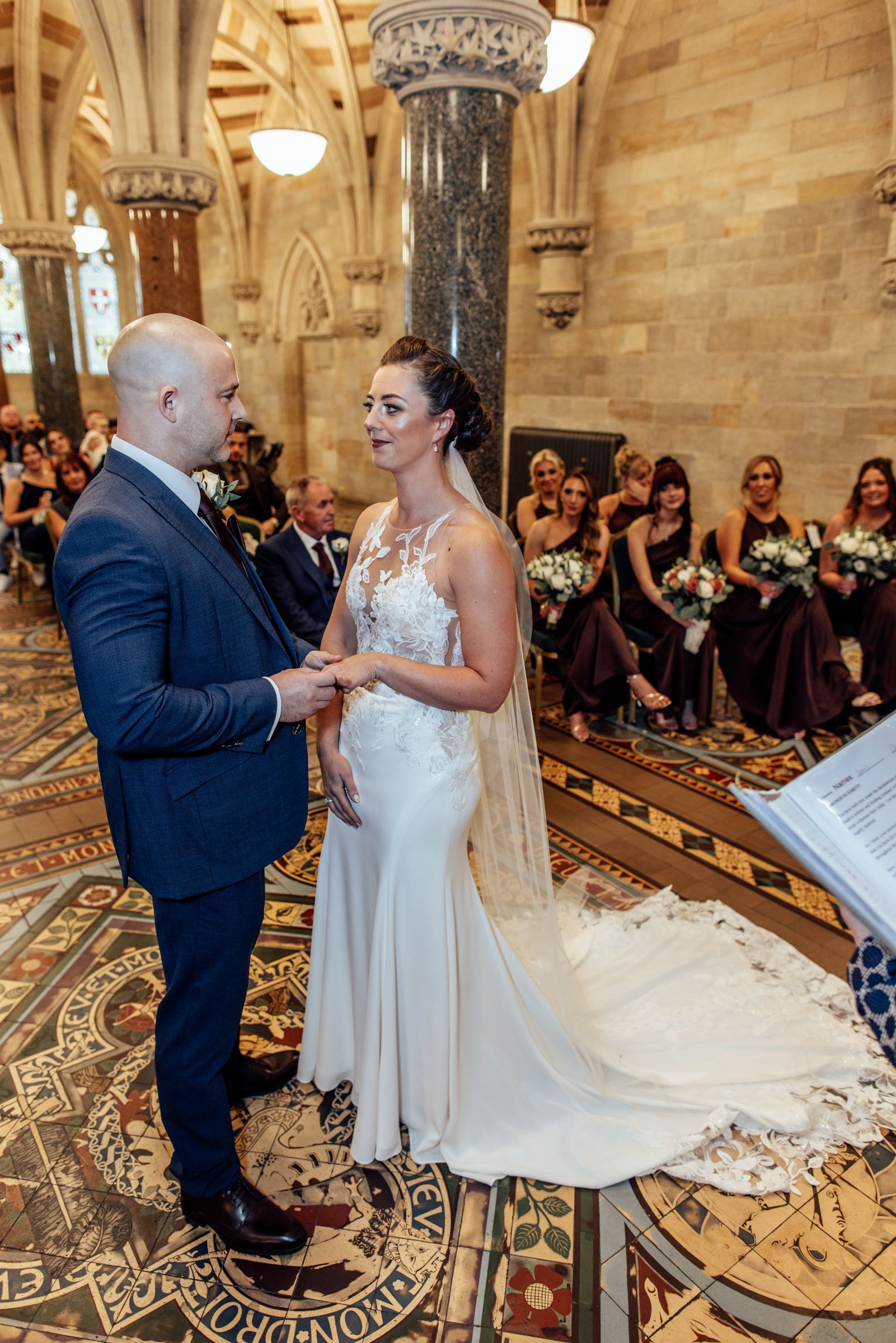 Bride in a lace gown and groom in a navy suit exchange vows in an ornate stone church as bridesmaids and guests look on.