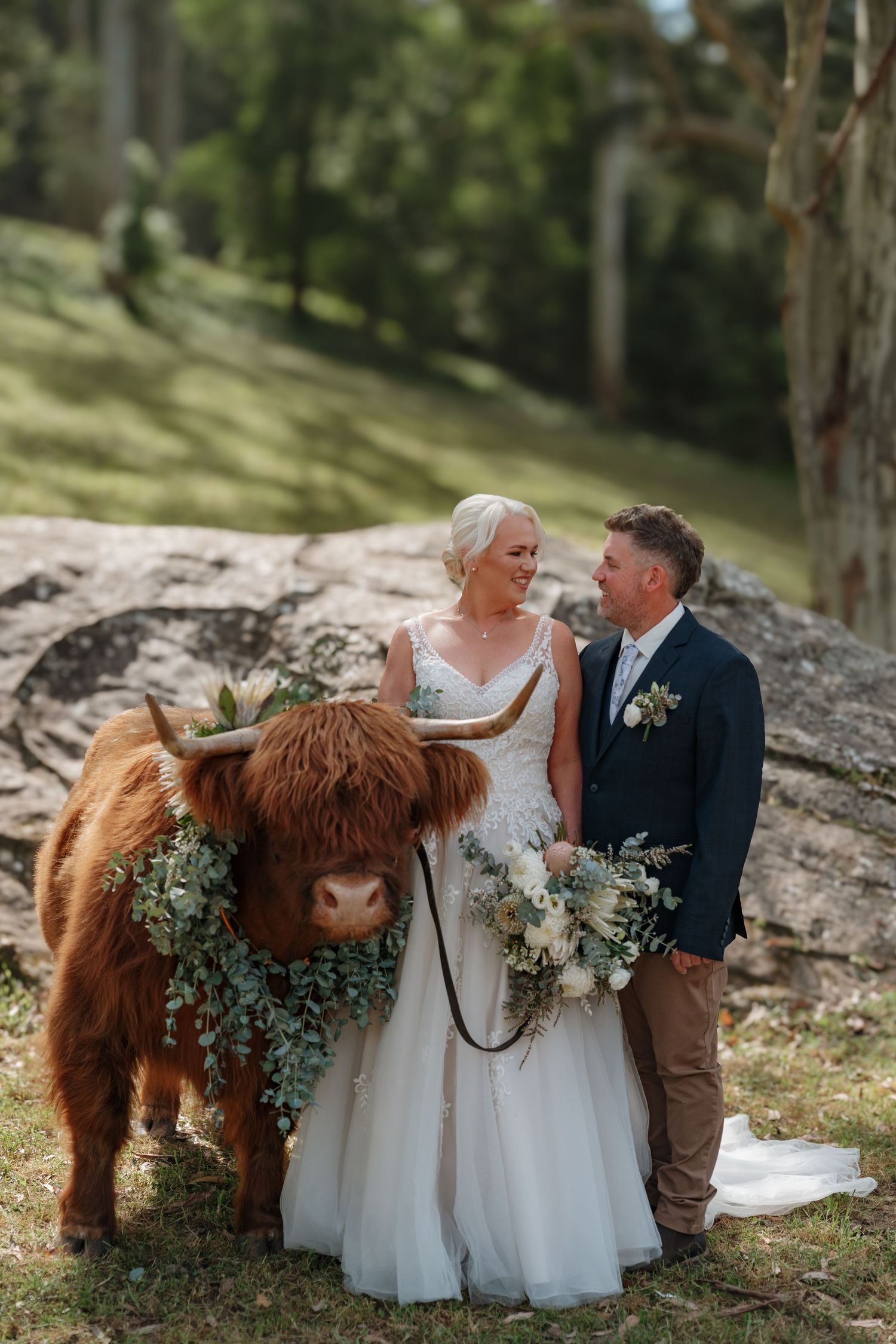 Bride and groom pose with a floral-adorned Highland cow at an outdoor rustic wedding ceremony.