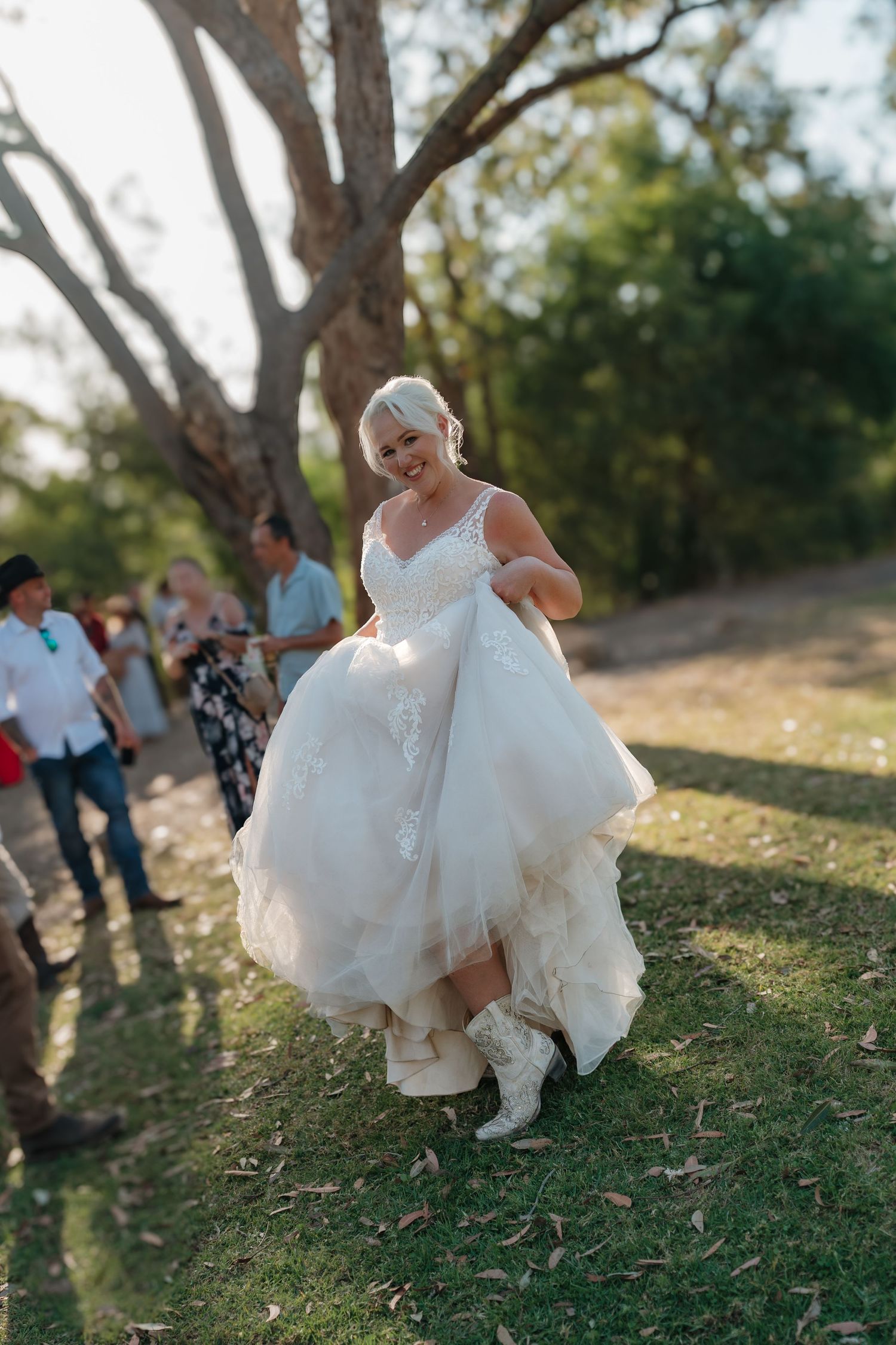 Smiling bride in white wedding gown and cowboy boots lifts her dress while walking outdoors at a rustic wedding celebration.