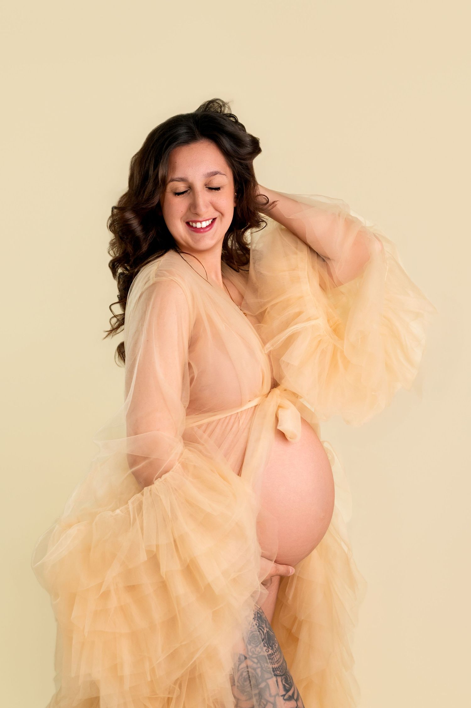 Smiling pregnant woman in sheer peach tulle robe posing against a soft yellow background, showcasing her baby bump.