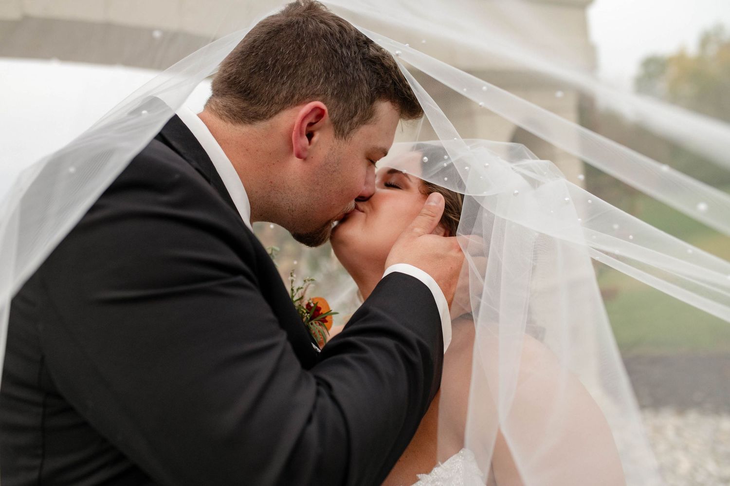 Bride and groom sharing a romantic kiss under a flowing white veil outdoors on their wedding day.