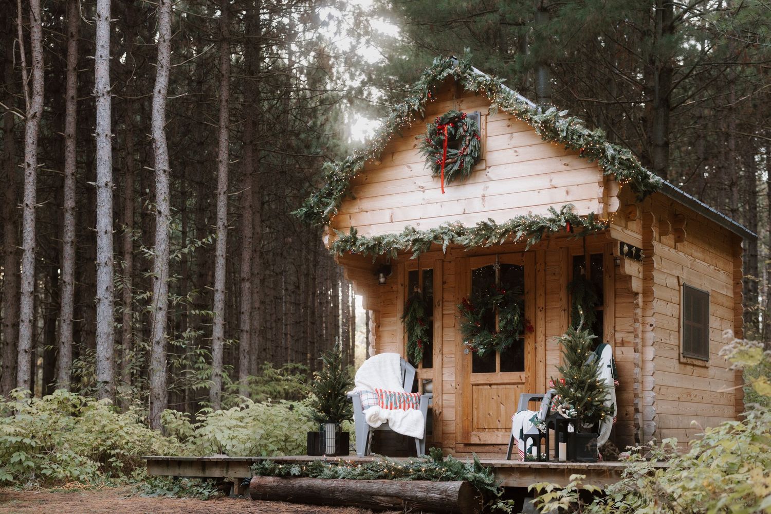 Cozy wooden cabin decorated with Christmas wreaths and garland nestled among tall pine trees in a forest setting.