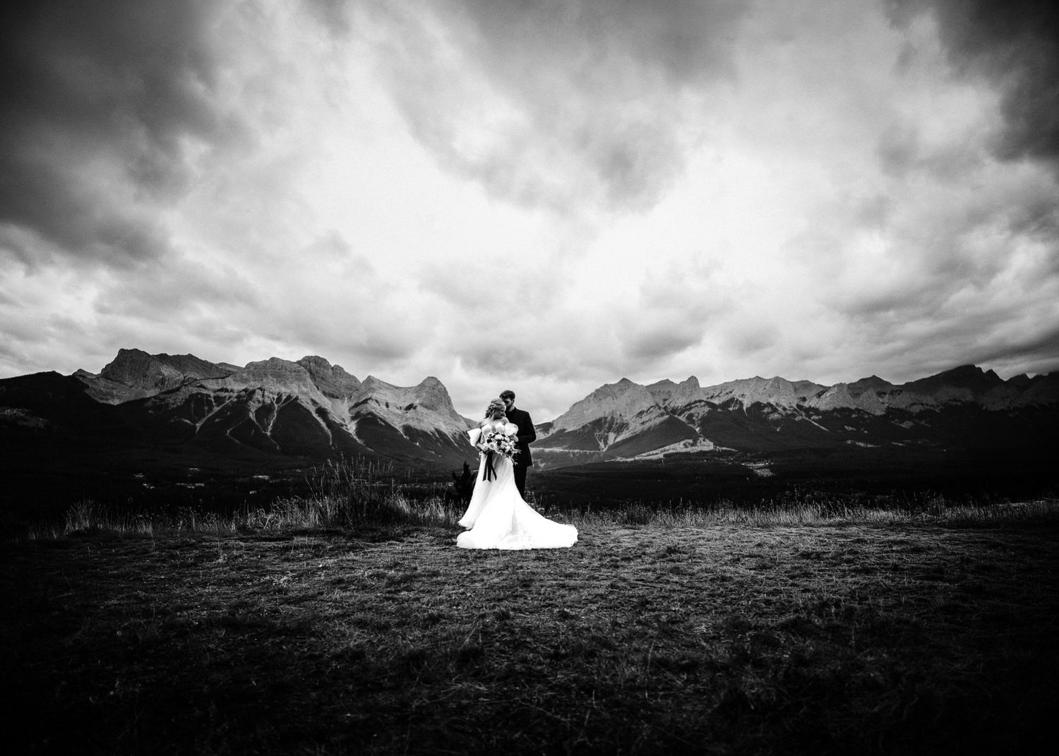 Black-and-white Canmore wedding photo of the couple in a wide field with mountains and dramatic clouds