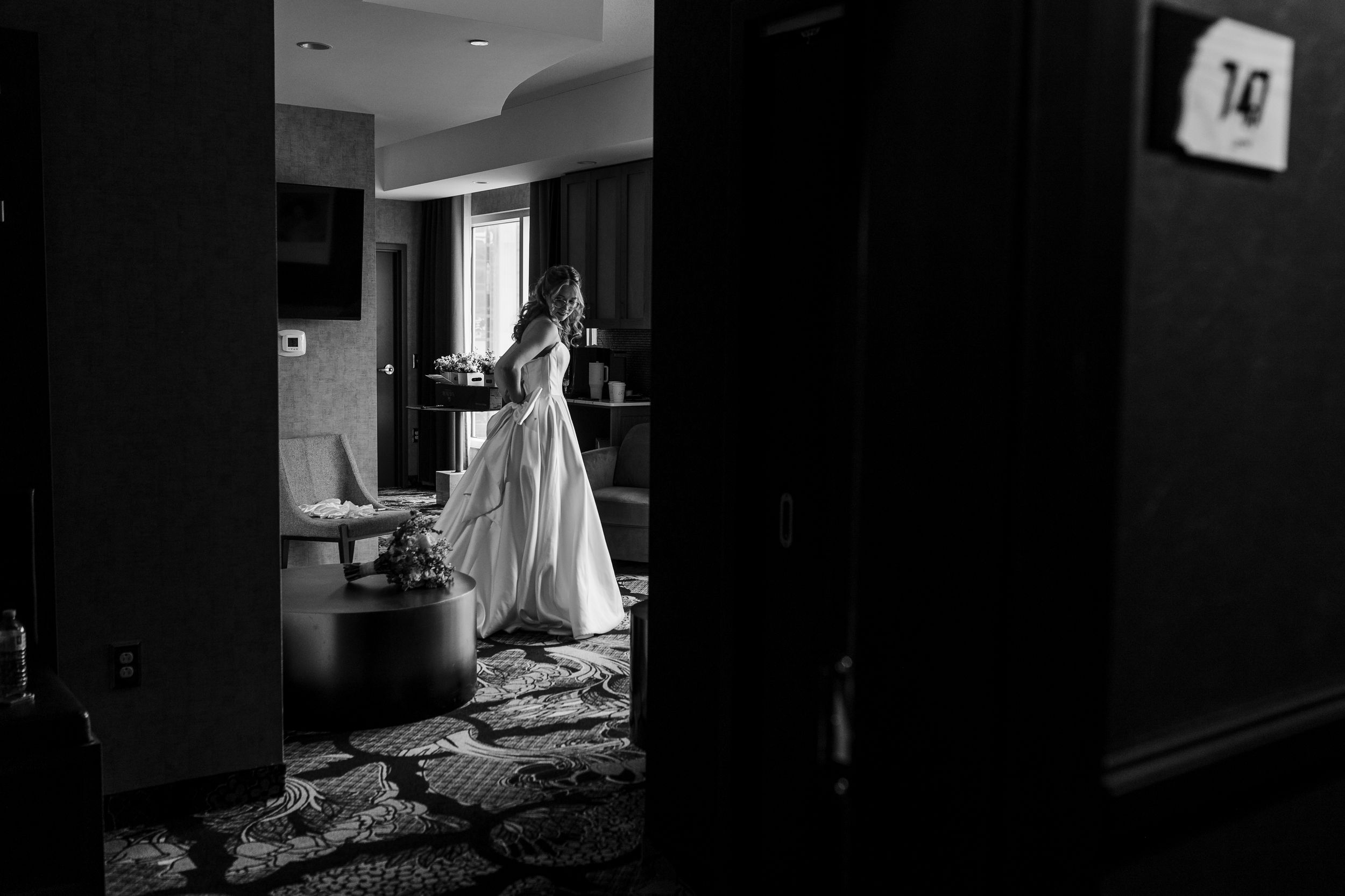 Black and white photo of a bride in a wedding gown standing in a hotel room doorway, illuminated by natural window light.