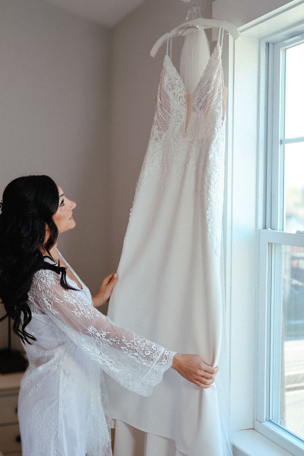 Bride in a lace robe admiring her wedding dress by the window while getting ready in New Jersey