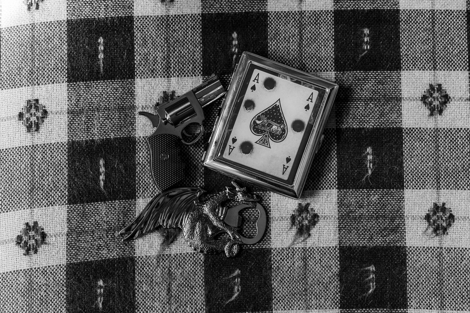 Black and white photo of a revolver, ace of spades card, and keys on a plaid cloth surface.
