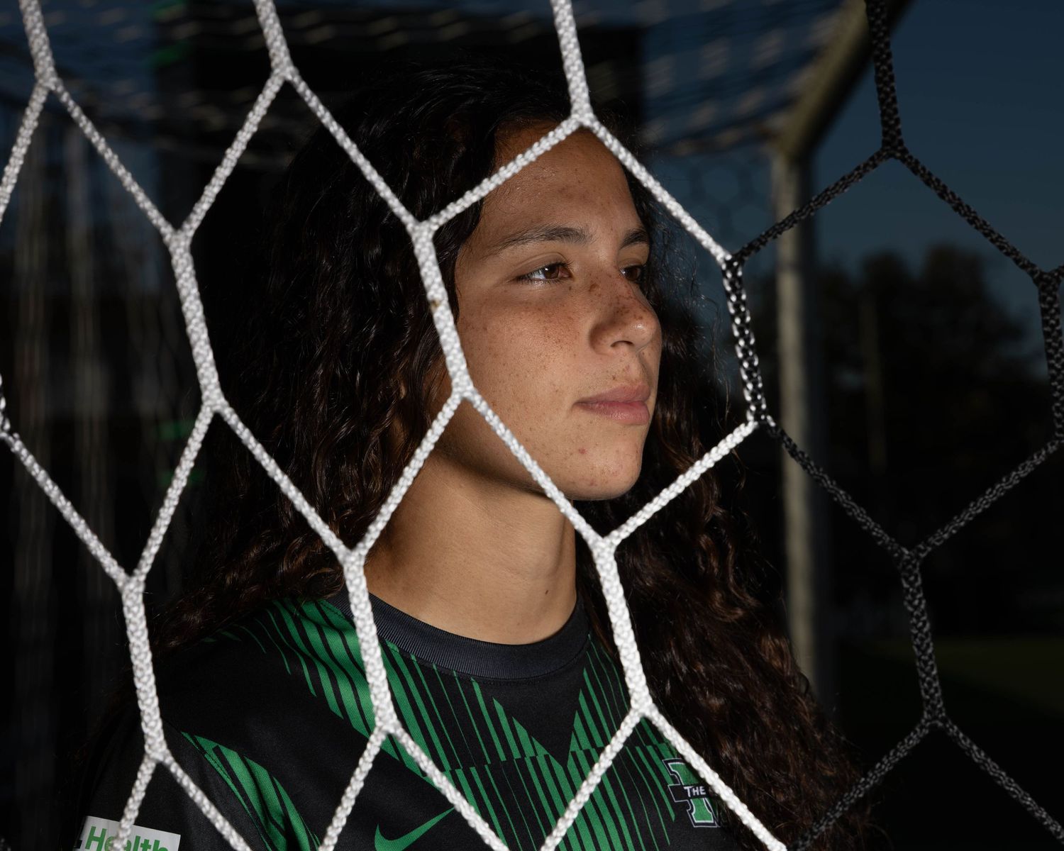 Female soccer goalkeeper poses behind a white net wearing a green and black Nike jersey at an outdoor field.