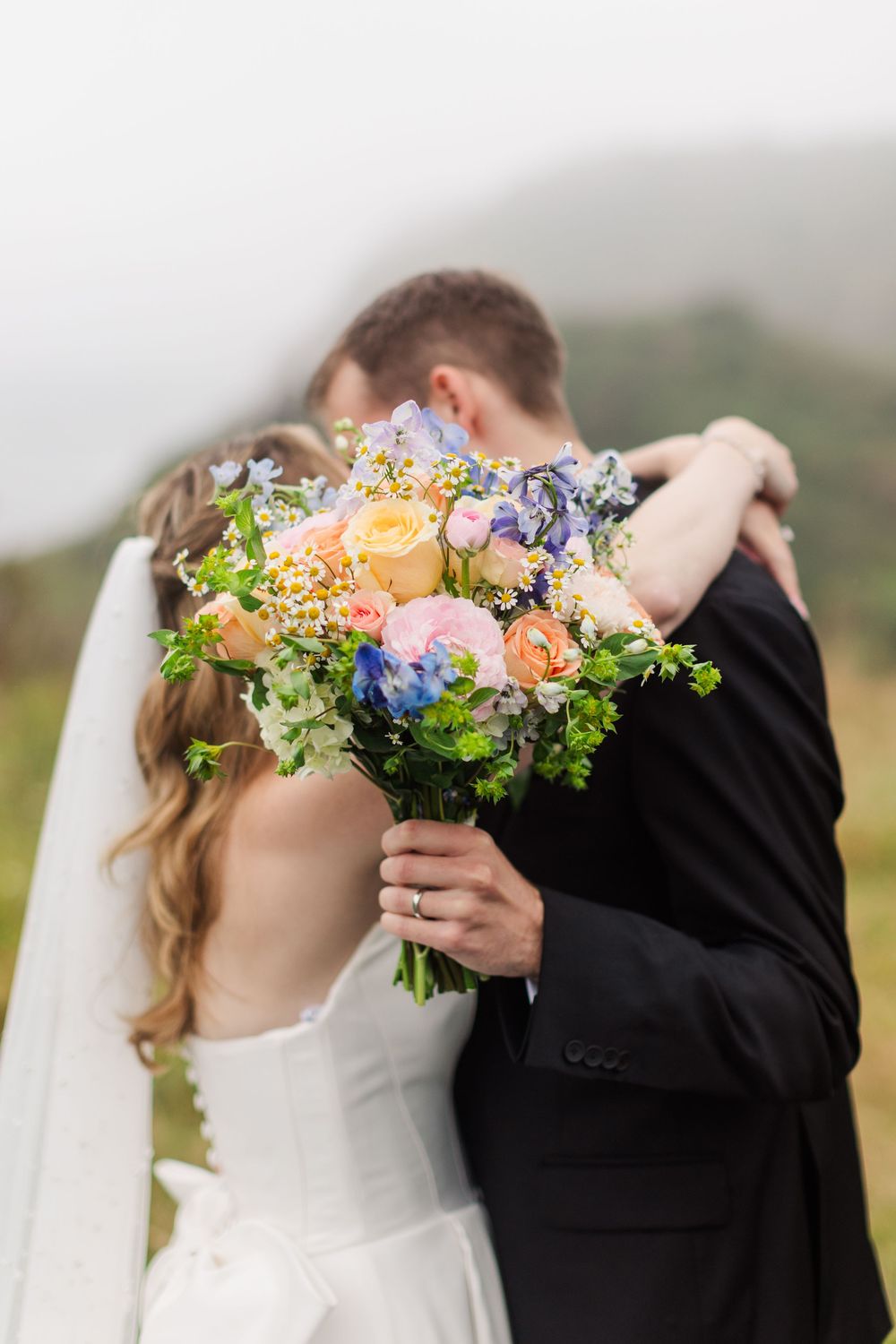 Bride and groom embracing outdoors holding a colorful spring wedding bouquet with blue, pink, and peach blooms.