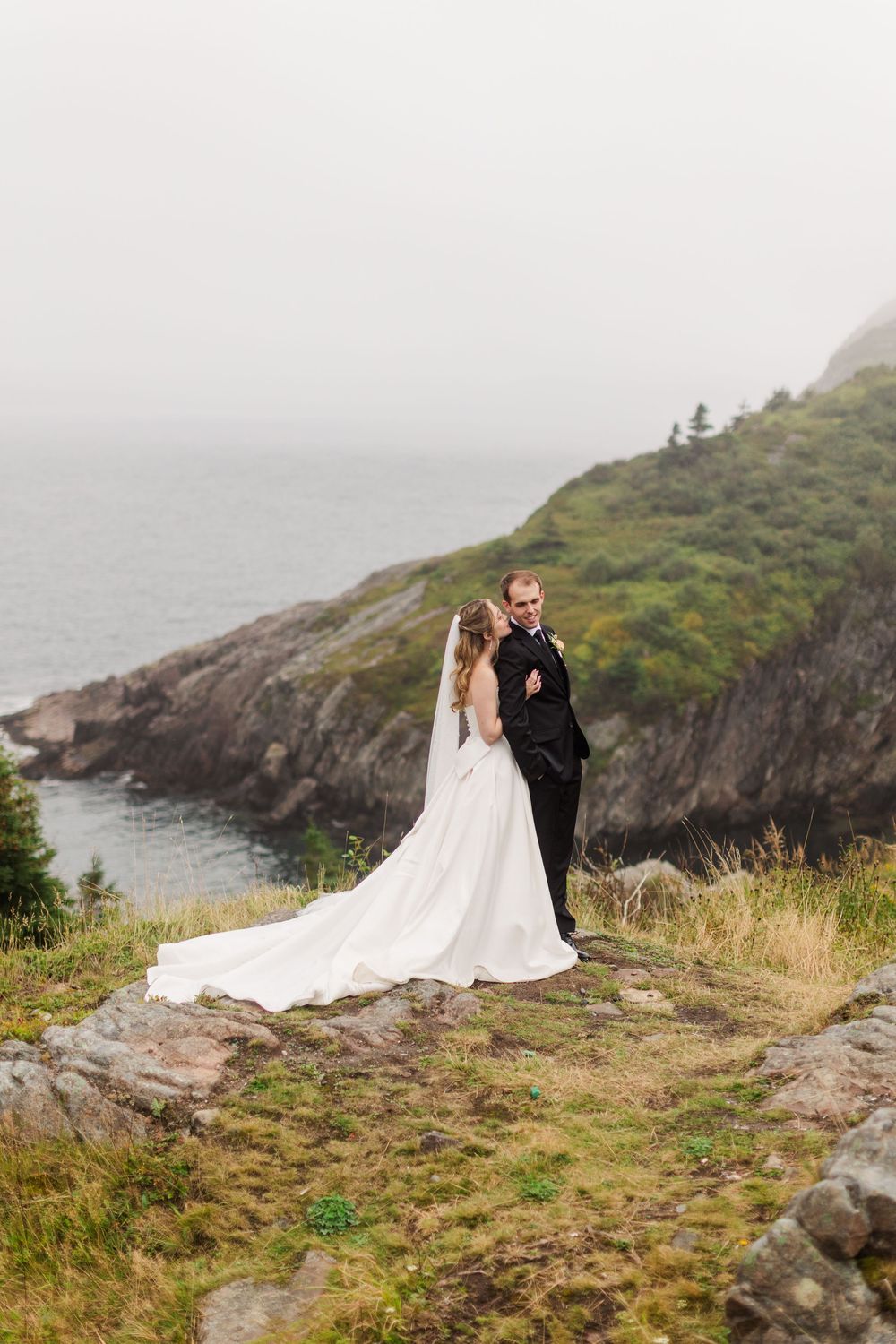 A bride and groom embrace on a rocky coastal cliff, her white gown trailing behind them with misty ocean views.